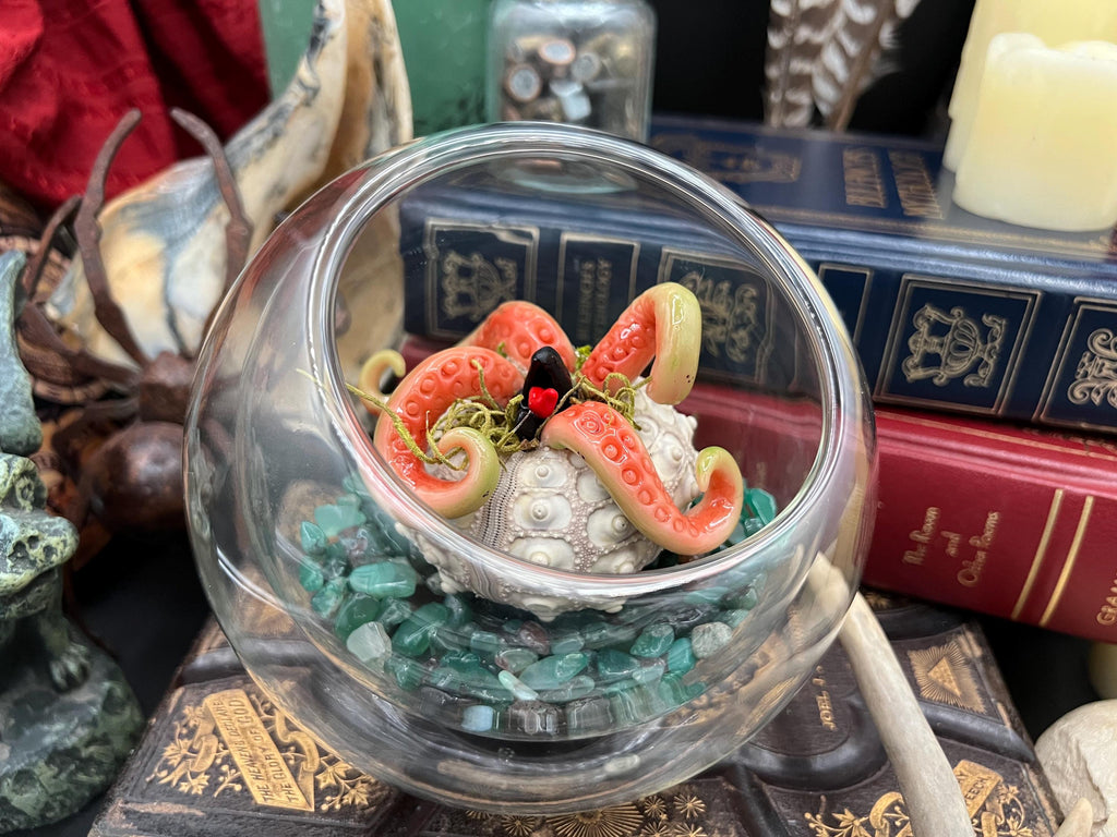 A decorative clear glass bowl filled with green stones and a small sculpted octopus figure with pink tentacles, displayed among books and other objects.