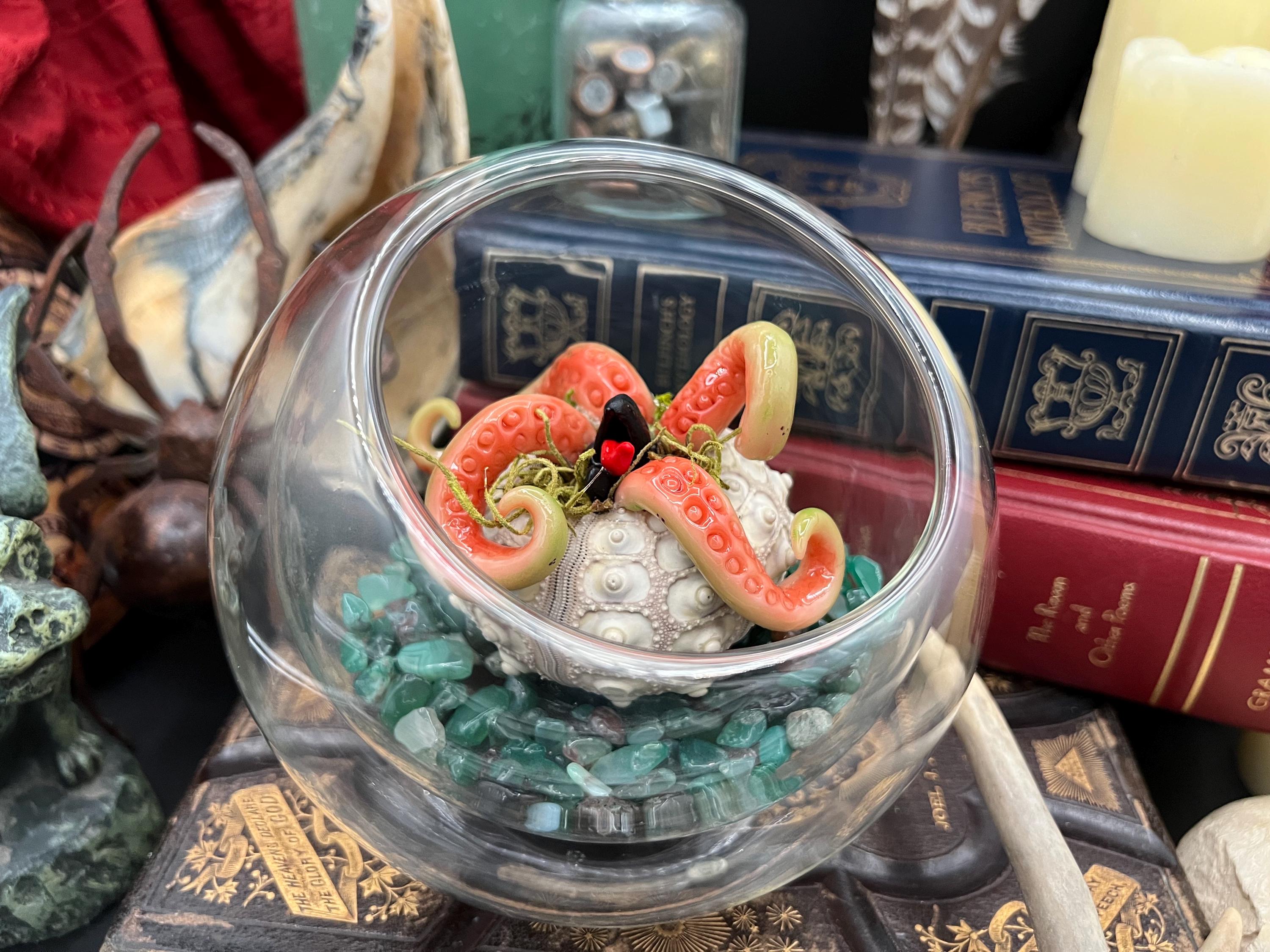 A decorative clear glass bowl filled with green stones and a small sculpted octopus figure with pink tentacles, displayed among books and other objects.