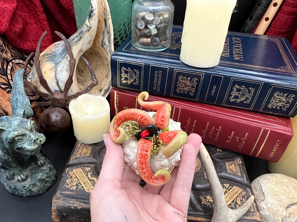 A hand holds a small decorative figurine shaped like a coral or sea-creature with orange-red tentacles, pale beige rock-like body sections, and small dark red and gold details, surrounded by books, candles, and other decorative objects.