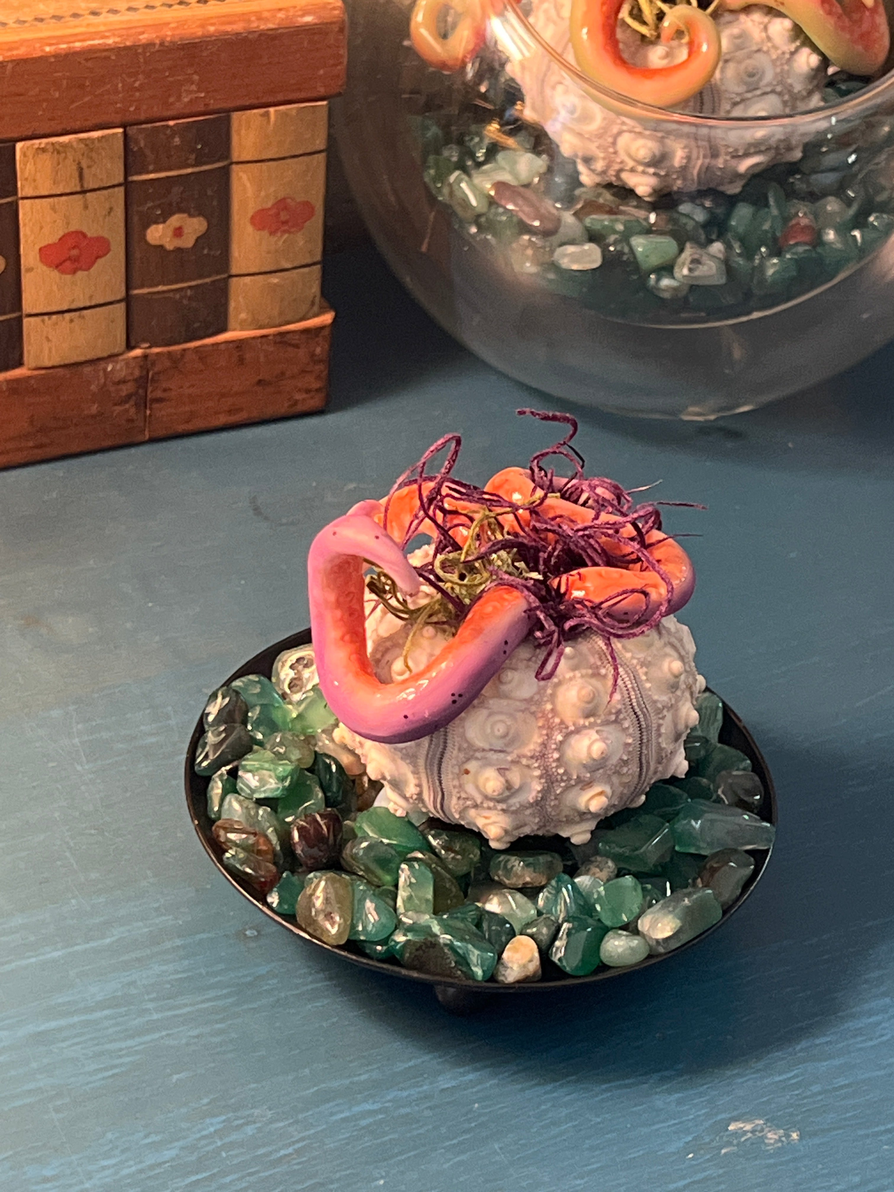 A decorative miniature figurine of a pink tentacula-like creature with purple tentacles, mounted on a gray bivalve shell and surrounded by green polished stones, displayed on a blue surface.