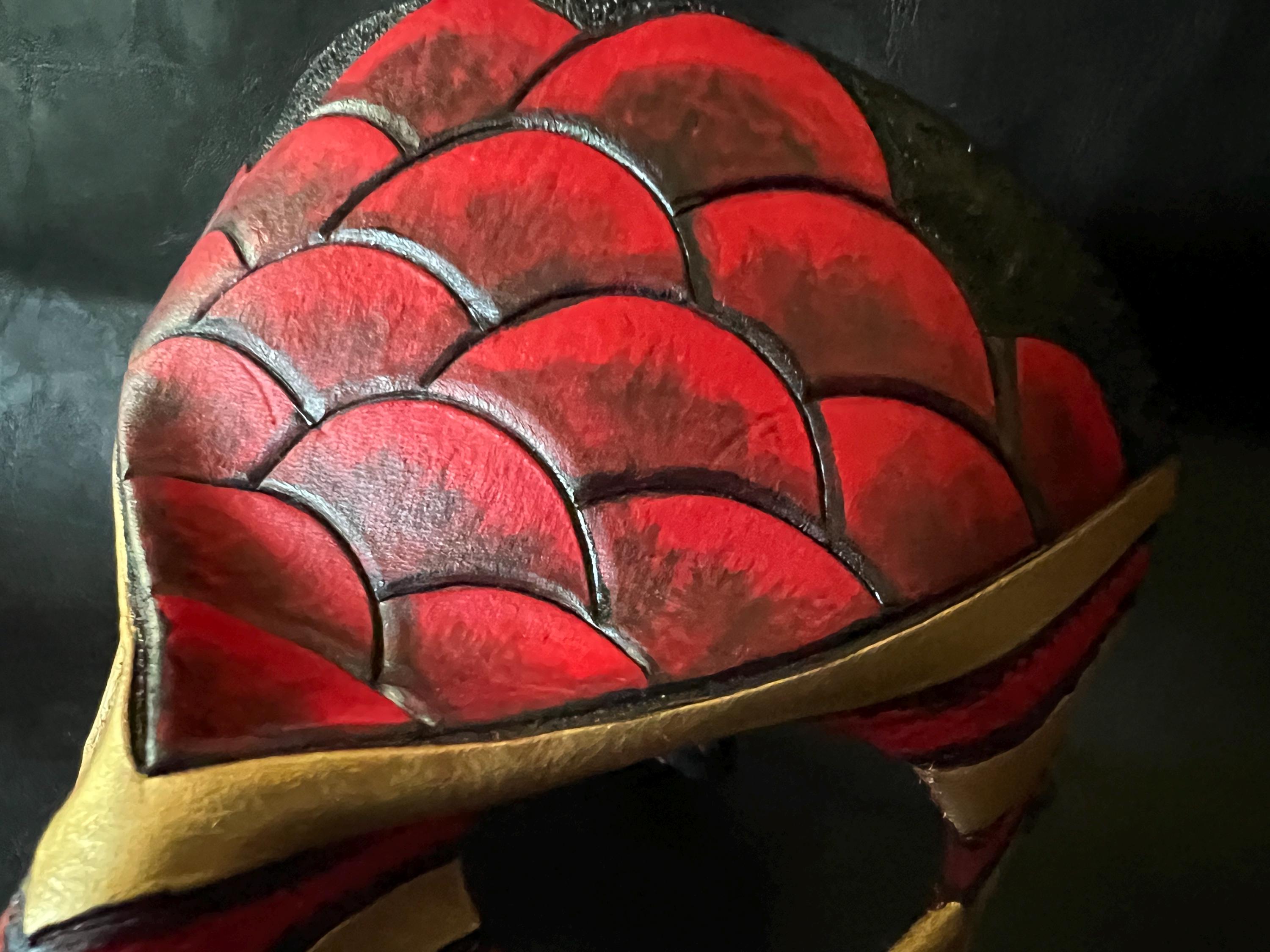A close-up of a red and black scaly armor piece with gold trim, showing detailed embossed scales and a curved gold border against a dark background.