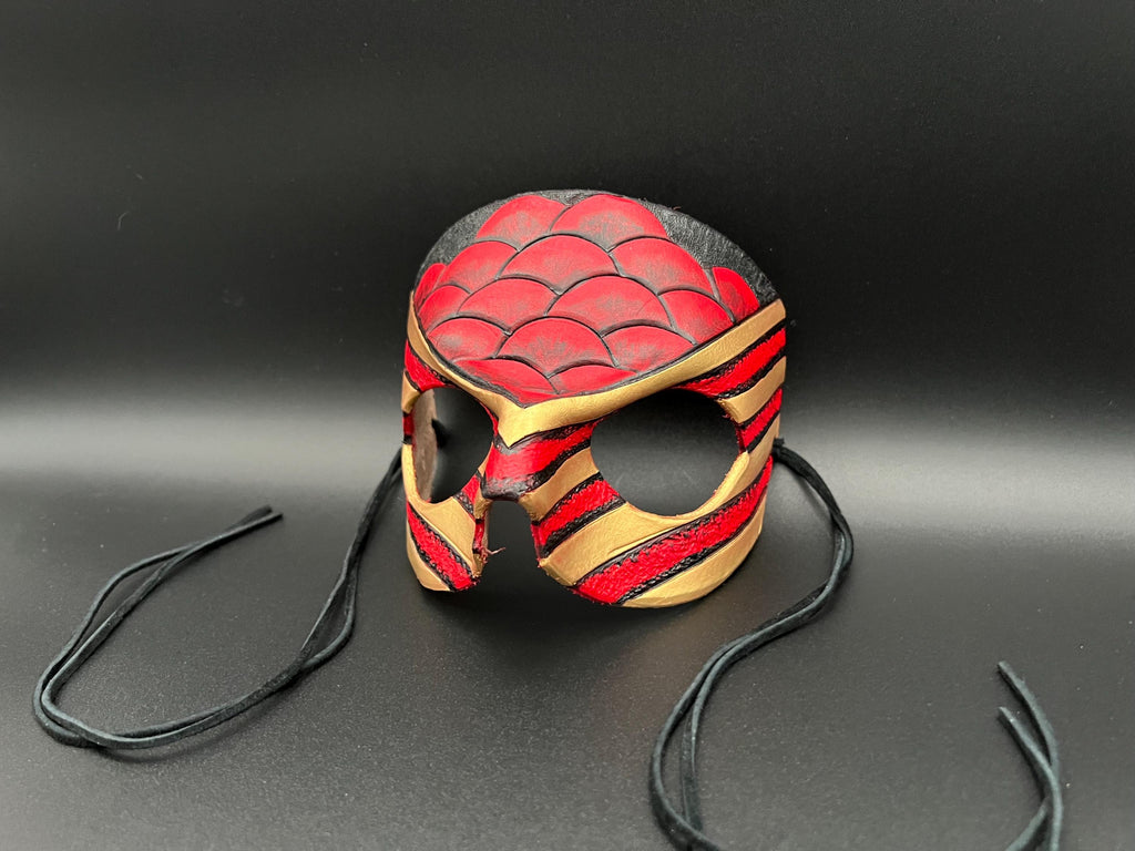 A handcrafted leather mask styled like a superhero or dragon face, featuring a red and black scaled pattern on the forehead, red and gold stripes on the cheeks, and black ear loops, set against a dark gray background.