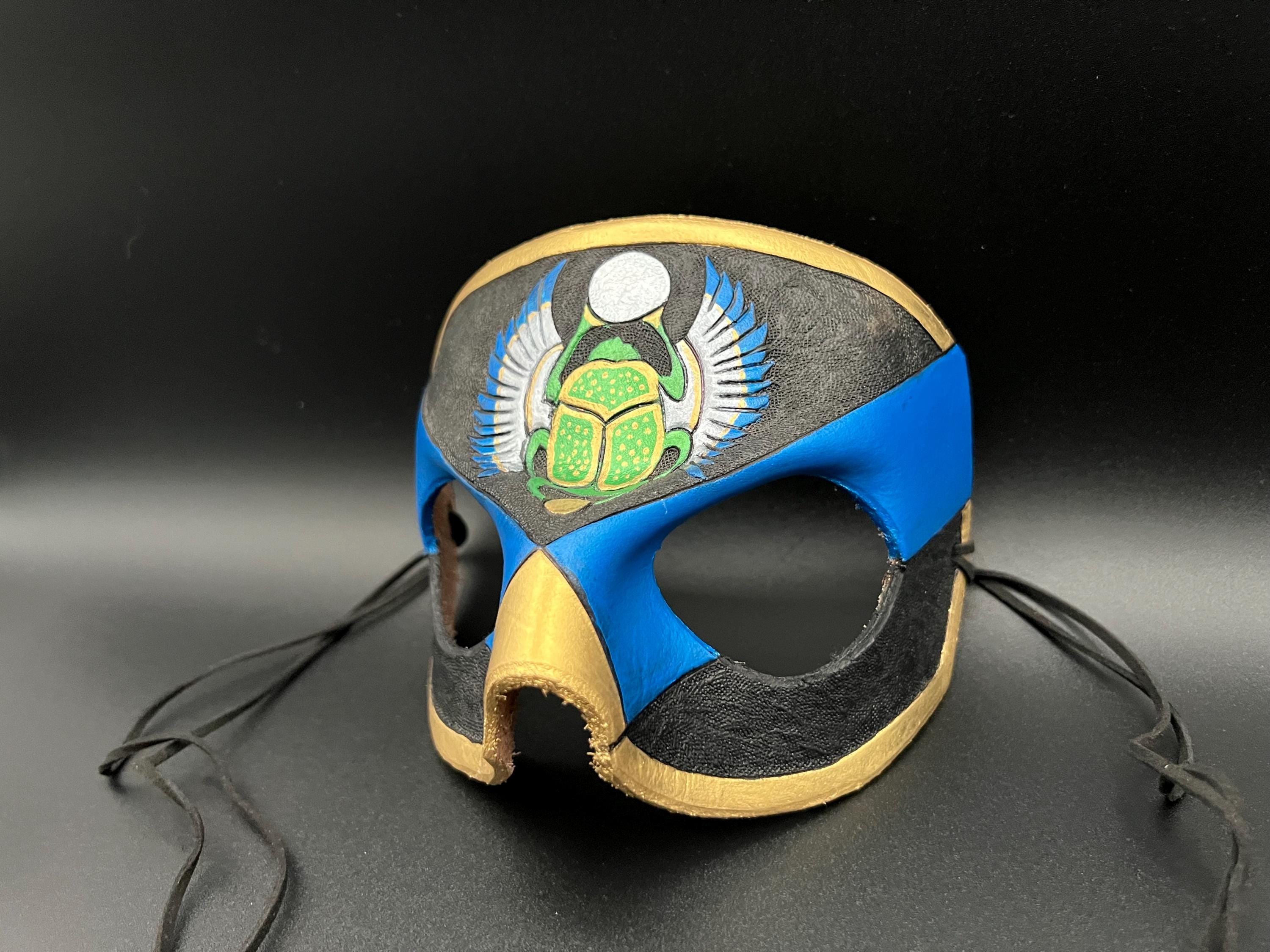 A leather mask featuring a blue and black design with gold trim, a green scarab beetle motif, and silver wings, shown on a dark gray background with black ties