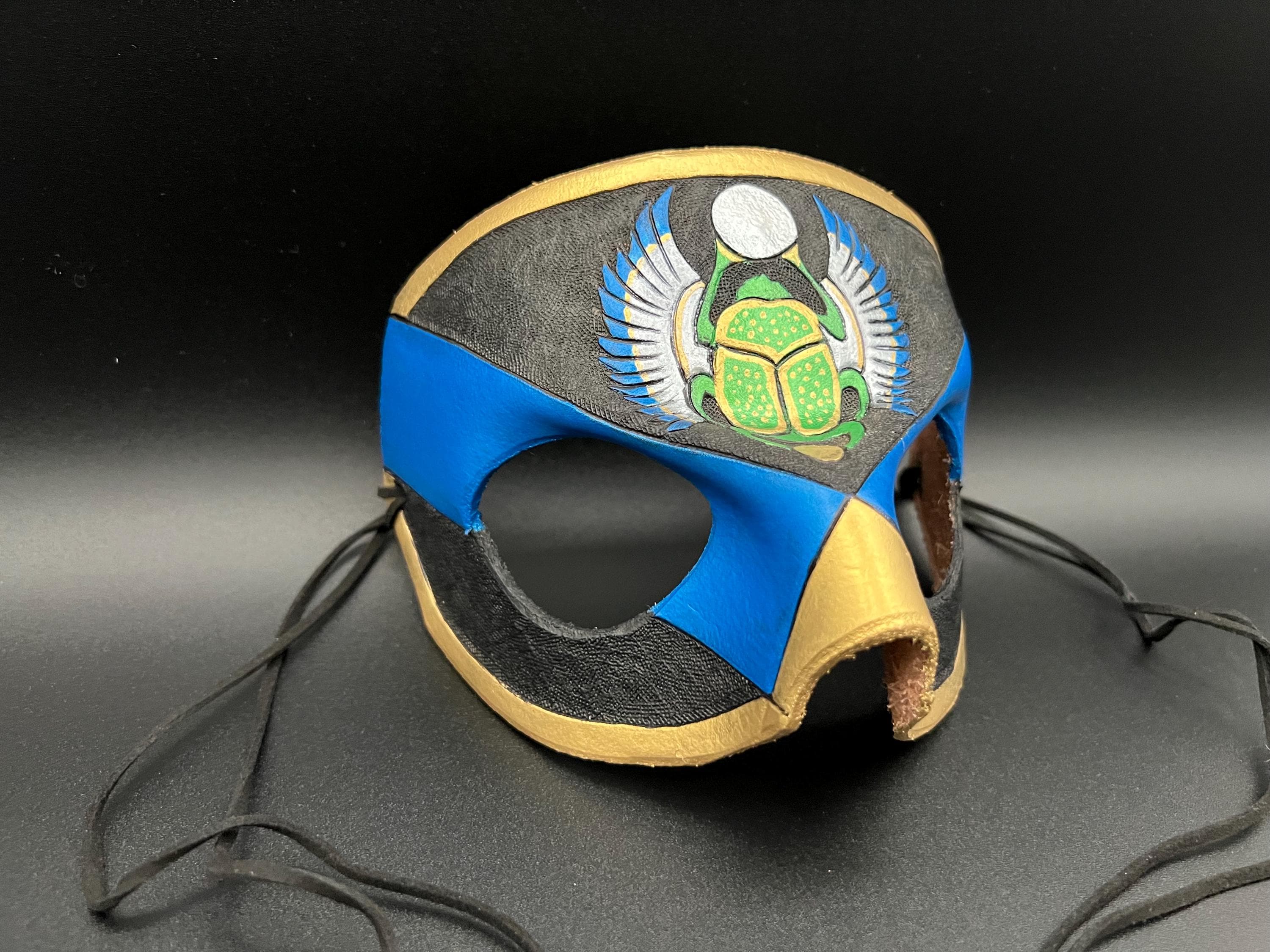 A handcrafted leather mask featuring a blue and gold design with a painted green scarab beetle motif on the forehead, displayed against a dark background.