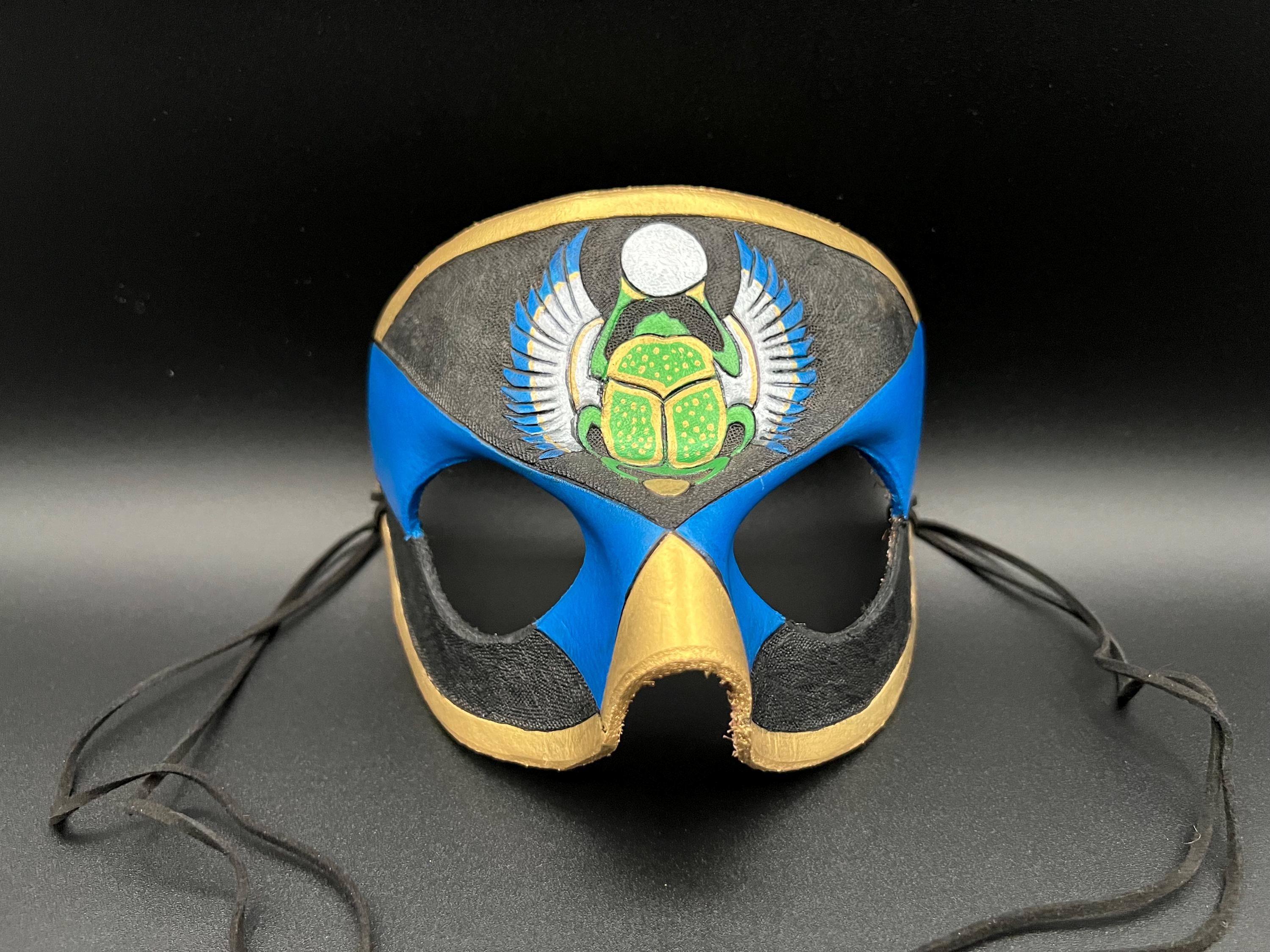 A leather mask featuring a blue and black design with gold trim, a green scarab beetle motif, and silver wings, shown on a dark gray background with black ties