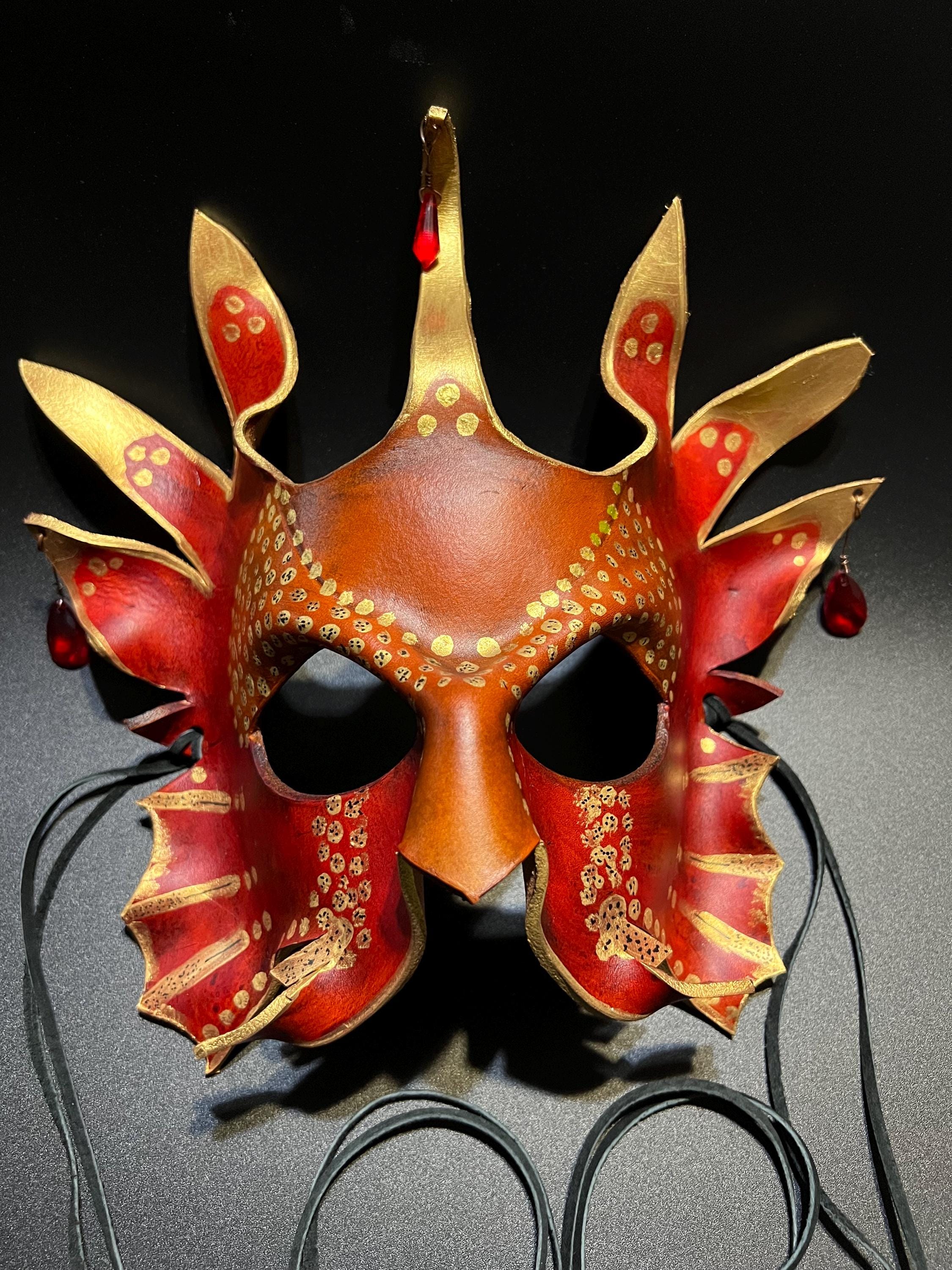 A decorative masquerade mask with a red and gold dragon-inspired design, featuring pointed horns, eye cutouts, and dangling red beads, displayed against a dark background.
