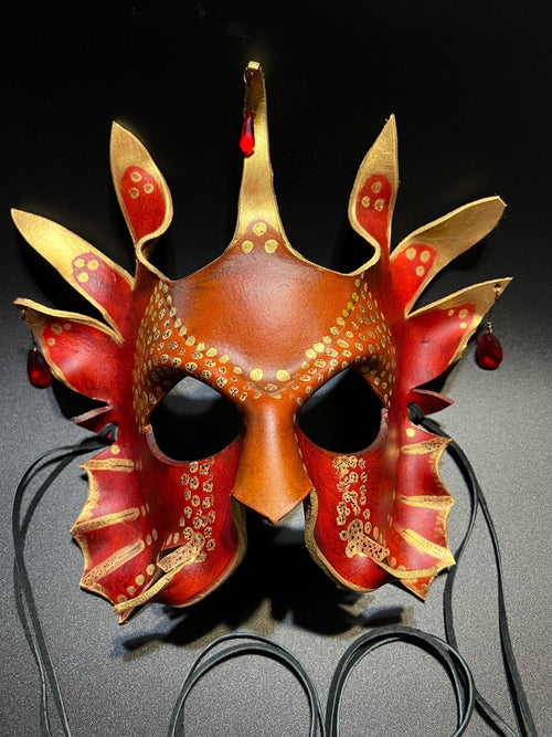 A decorative masquerade mask with a red and gold dragon-inspired design, featuring pointed horns, eye cutouts, and dangling red beads, displayed against a dark background.