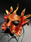 A decorative red and gold masquerade mask with multiple pointed flame-like extensions, gold dot patterns, and small red hanging beads, photographed on a dark background with black cords visible below.