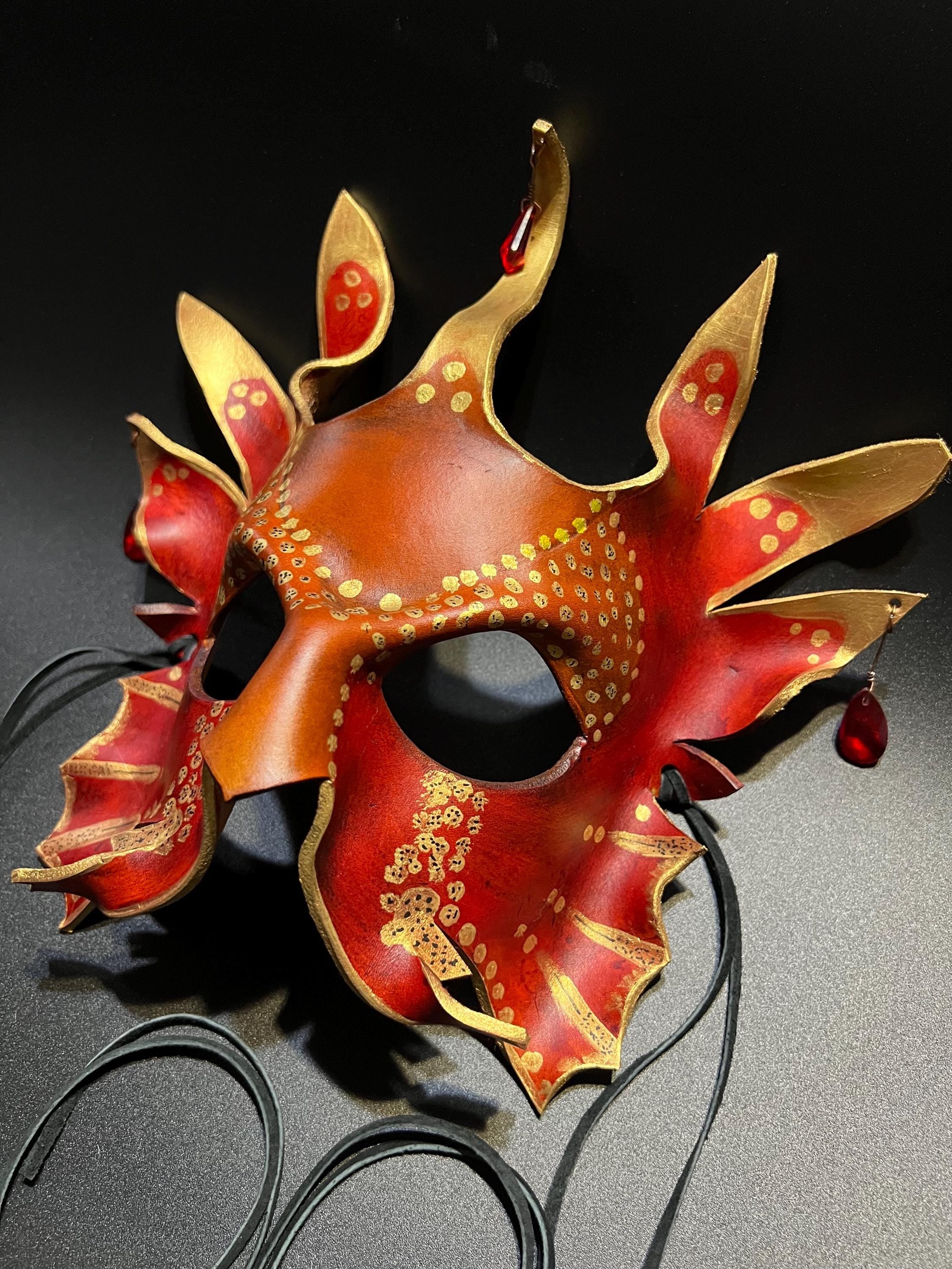 A decorative red and gold masquerade mask with multiple pointed flame-like extensions, gold dot patterns, and small red hanging beads, photographed on a dark background with black cords visible below.