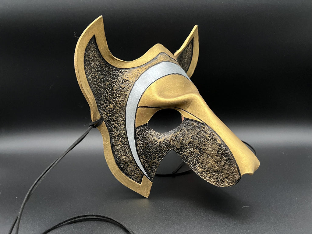 A stylized fantasy mask with a metallic gold and textured bronze finish, featuring a curved silver stripe and a black hanging cord, displayed against a dark background.