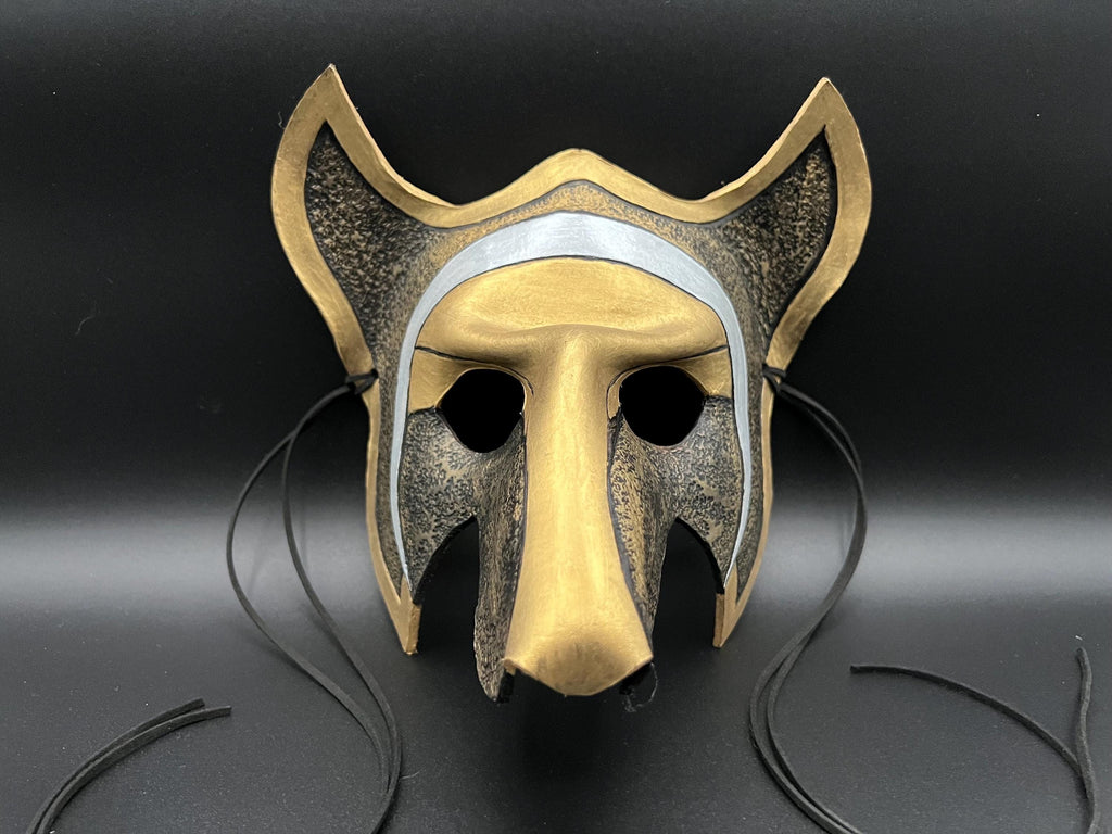 A stylized animal mask, resembling a fox or wolf, with a metallic gold finish, dark textured sides, a silver band, and black hanging ties, shown against a dark background.
