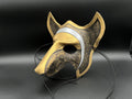 A stylized fox or wolf mask with a metallic gold finish, textured black and gold patterned sections, and a silver curved stripe, displayed on a black stand against a dark background.