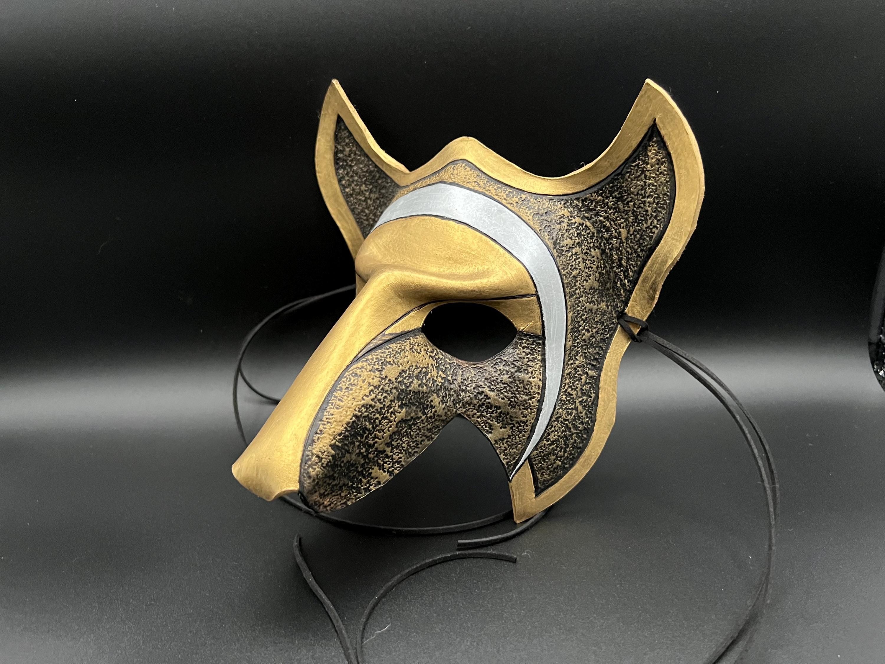A stylized fox or wolf mask with a metallic gold finish, textured black and gold patterned sections, and a silver curved stripe, displayed on a black stand against a dark background.
