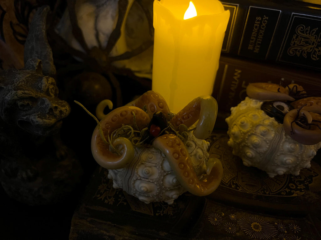 A gothic-style decorative set featuring two octopus-themed sculptures, one beige and one cream, arranged on a dark surface next to a glowing yellow pillar candle and old books.