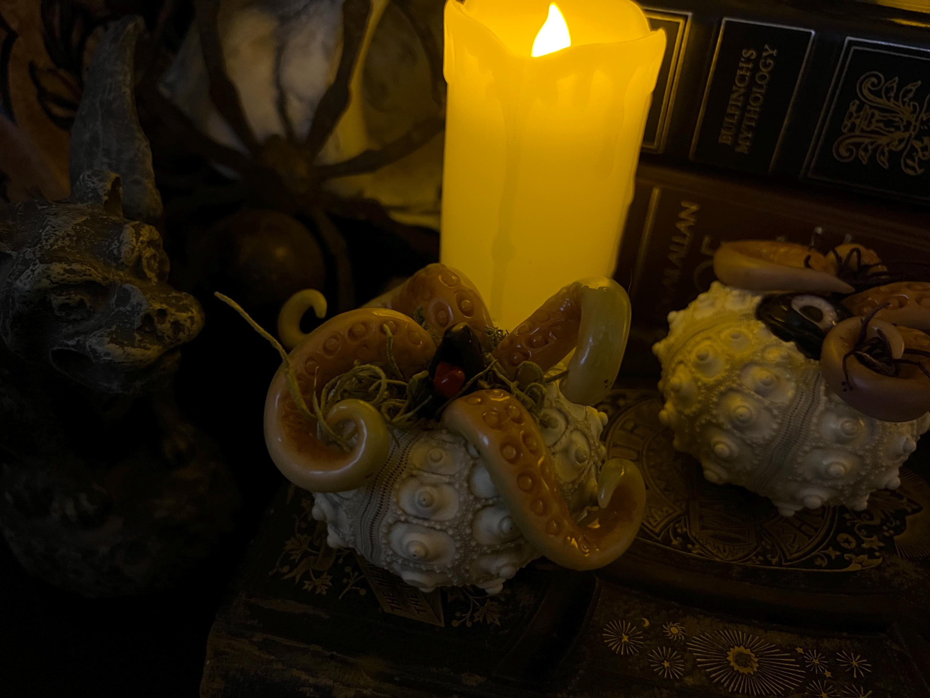 A gothic-style decorative set featuring two octopus-themed sculptures, one beige and one cream, arranged on a dark surface next to a glowing yellow pillar candle and old books.