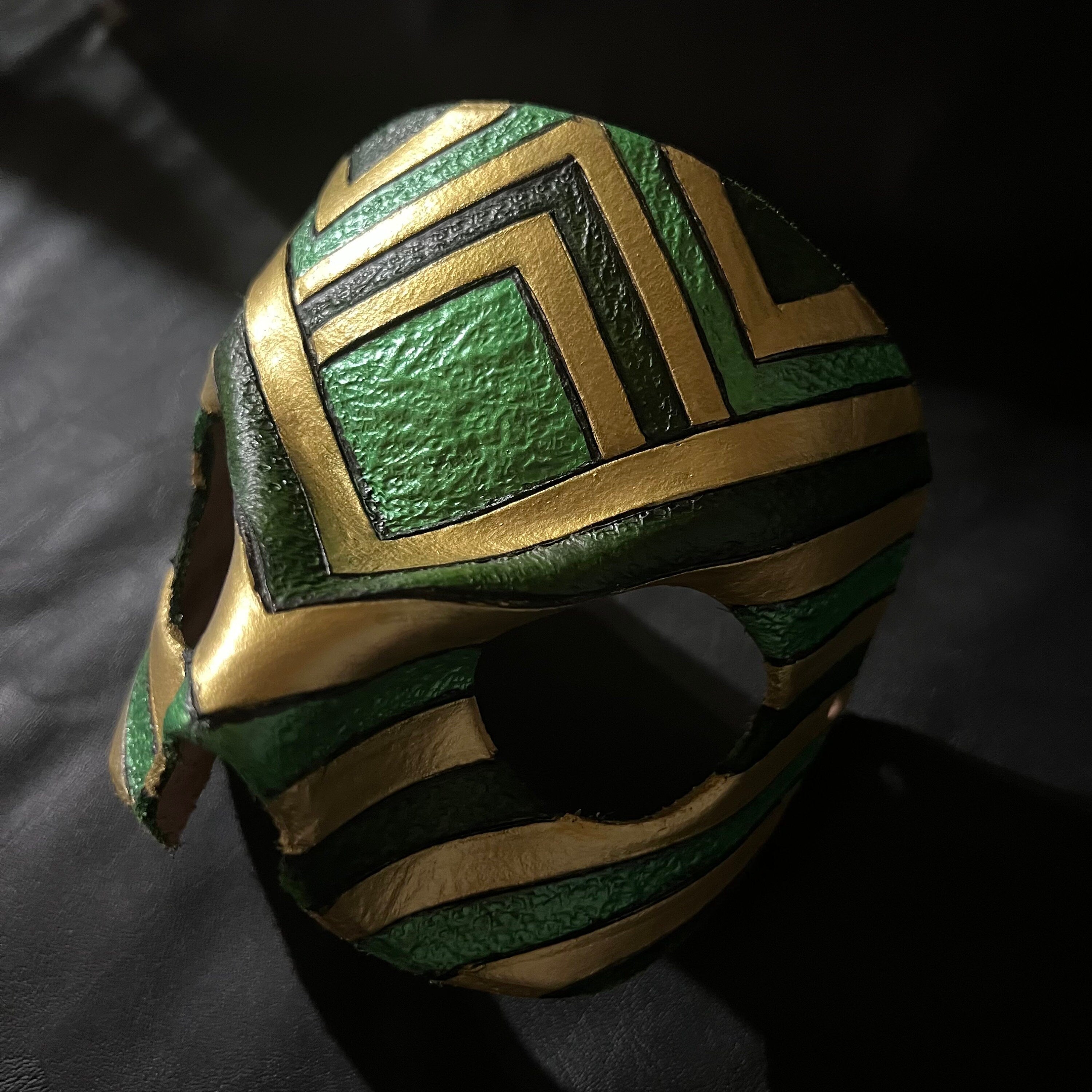 A close-up of a textured green and gold leather mask resting on a dark, textured surface.