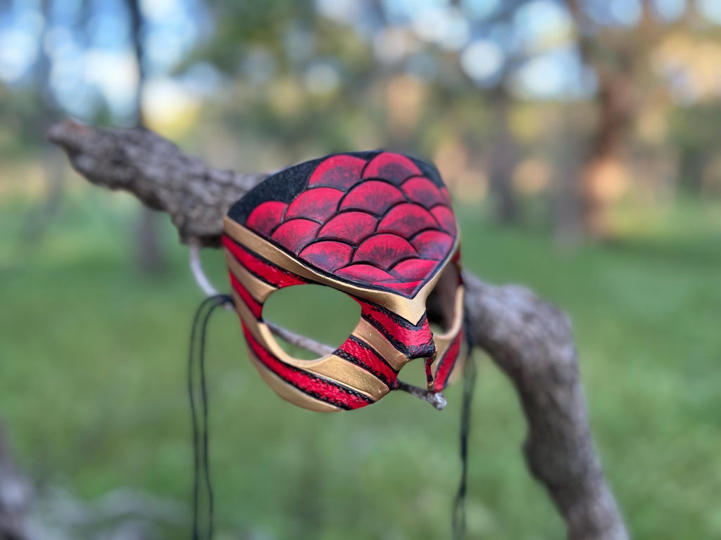 A handcrafted leather mask with a red and black scaled pattern and gold trim, hanging from a tree branch in an outdoor park setting.