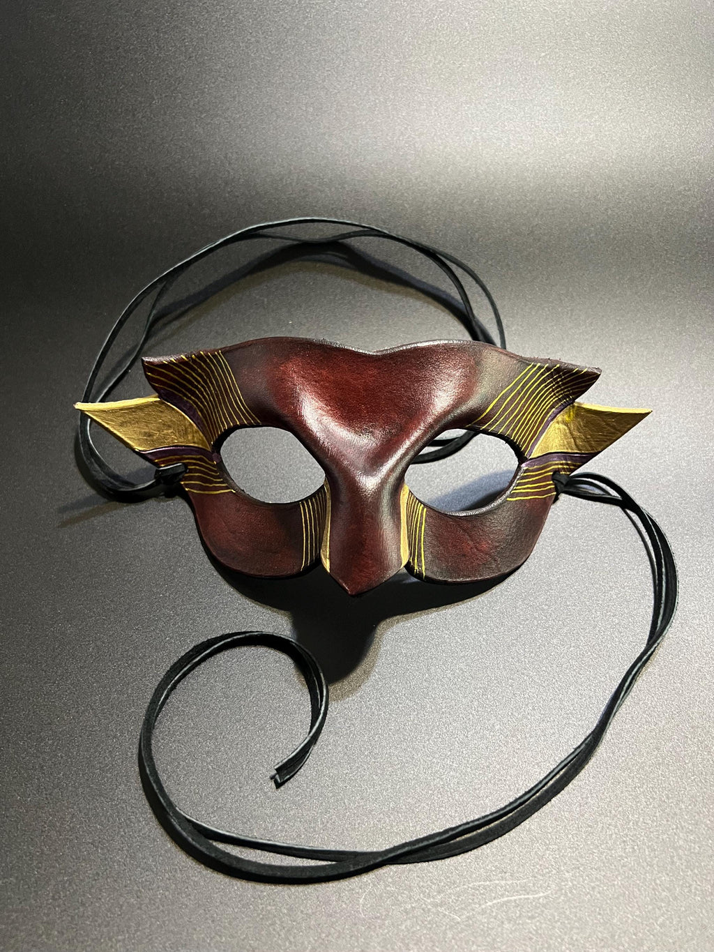 A decorative masquerade mask with a dark reddish-brown center, gold patterned side wings, and black suede ties, photographed on a gray studio background.