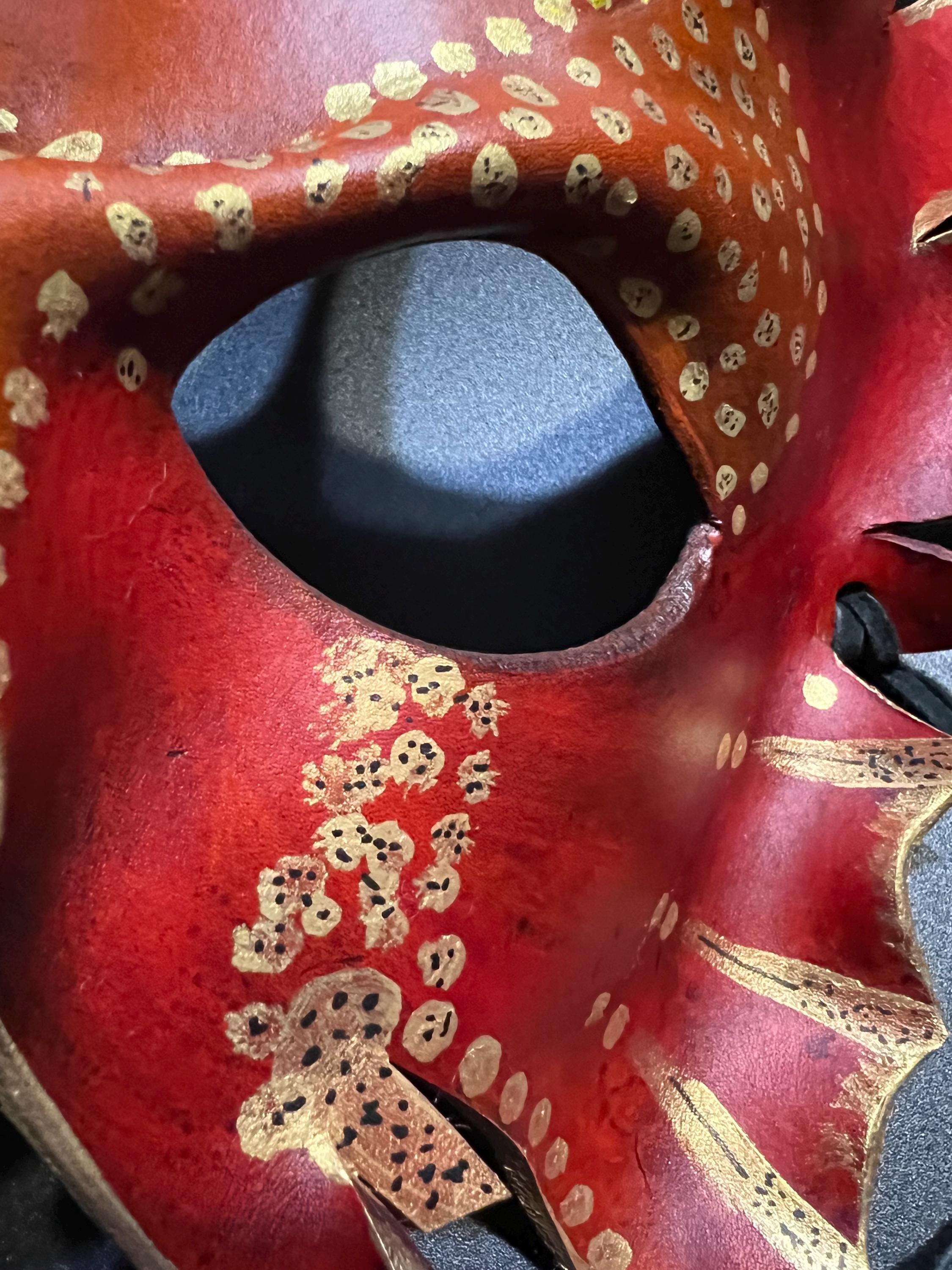 A close-up of a red masquerade mask with gold leaf accents and decorative spots, showing the eye opening and textured surface.