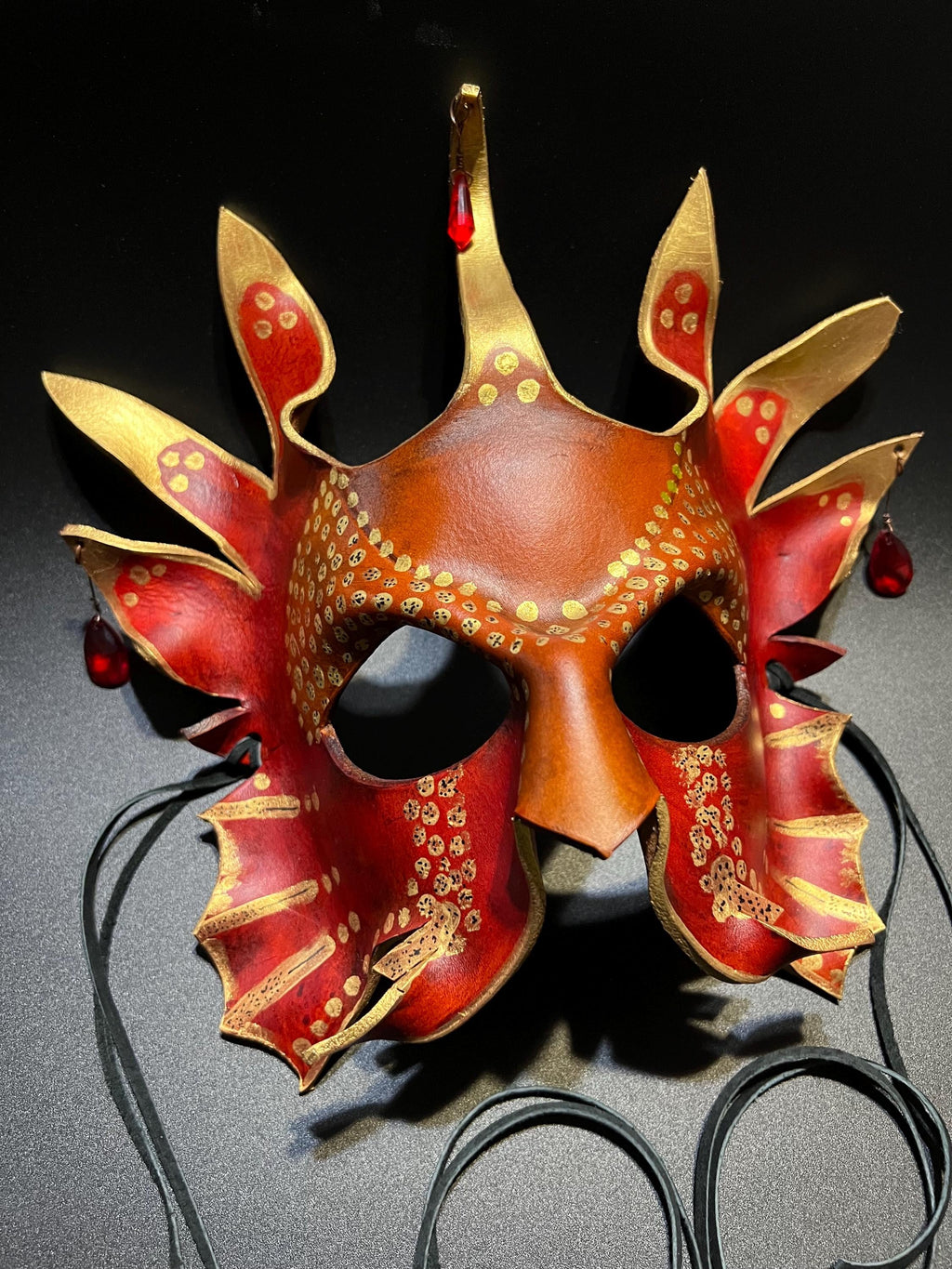 A decorative red and gold masquerade mask with pointed leaf-like extensions, gold painted details, and black hanging cords on a dark background.