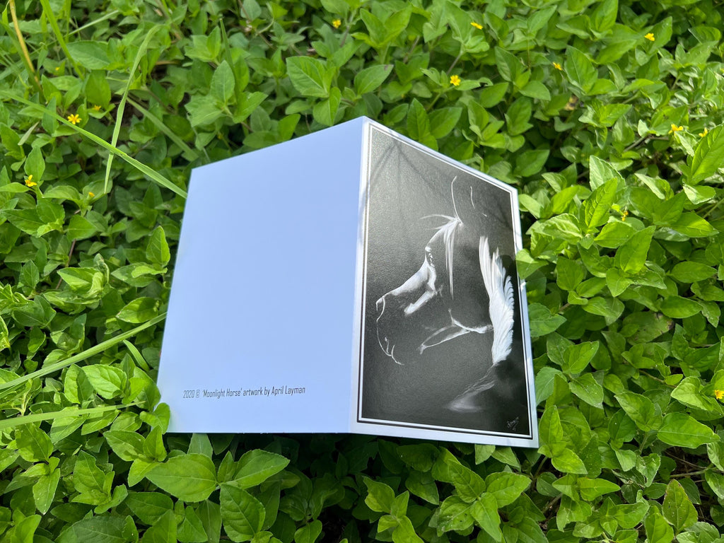 A square art print or card with a pale blue left panel and a black-and-white abstract figure print on the front, photographed outdoors on green foliage.