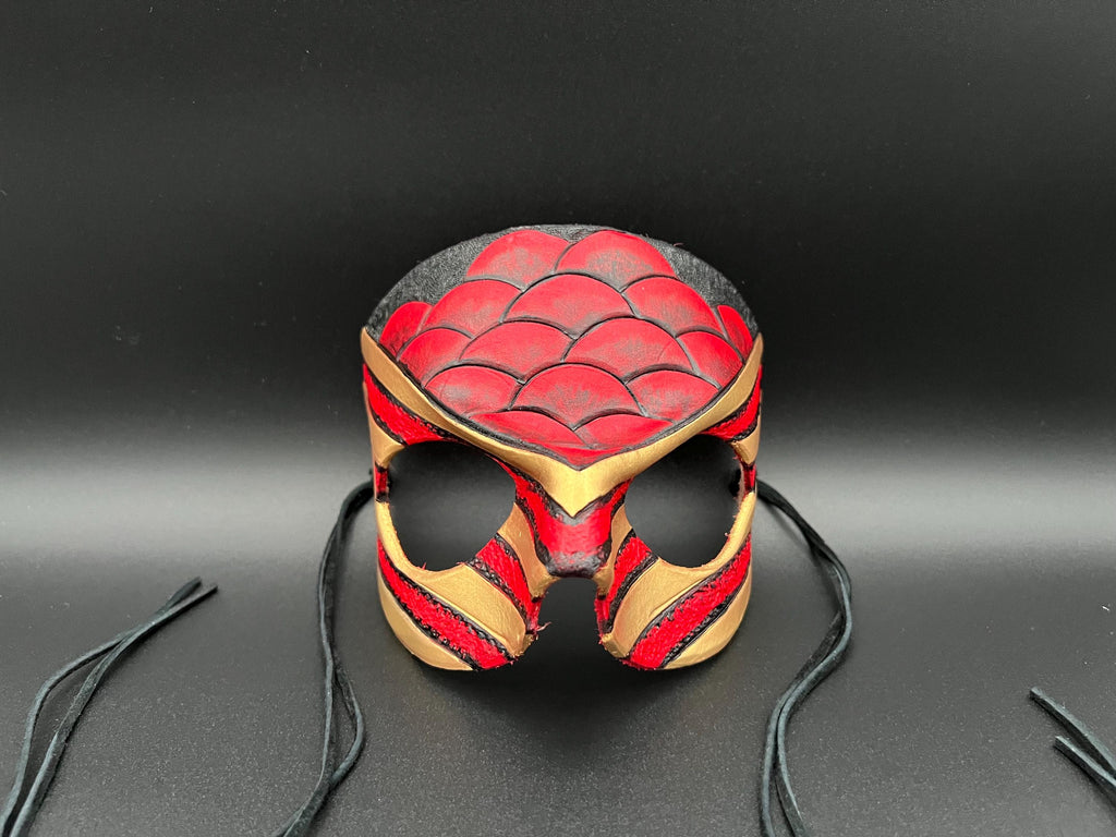 A leather mask featuring a red and black scales design with gold trim dragon motif shown on a dark gray background with black ties
