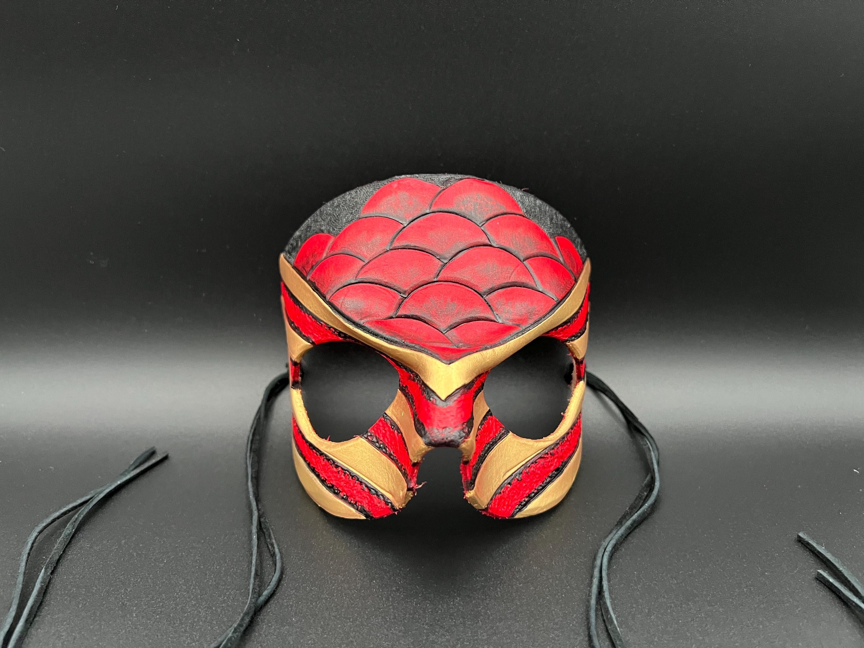 A leather mask featuring a red and black scales design with gold trim dragon motif shown on a dark gray background with black ties