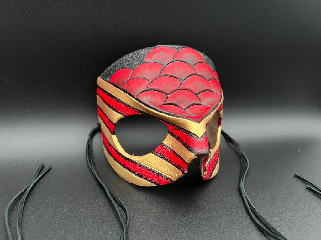 A leather mask featuring a red and black scales design with gold trim dragon motif shown on a dark gray background with black ties