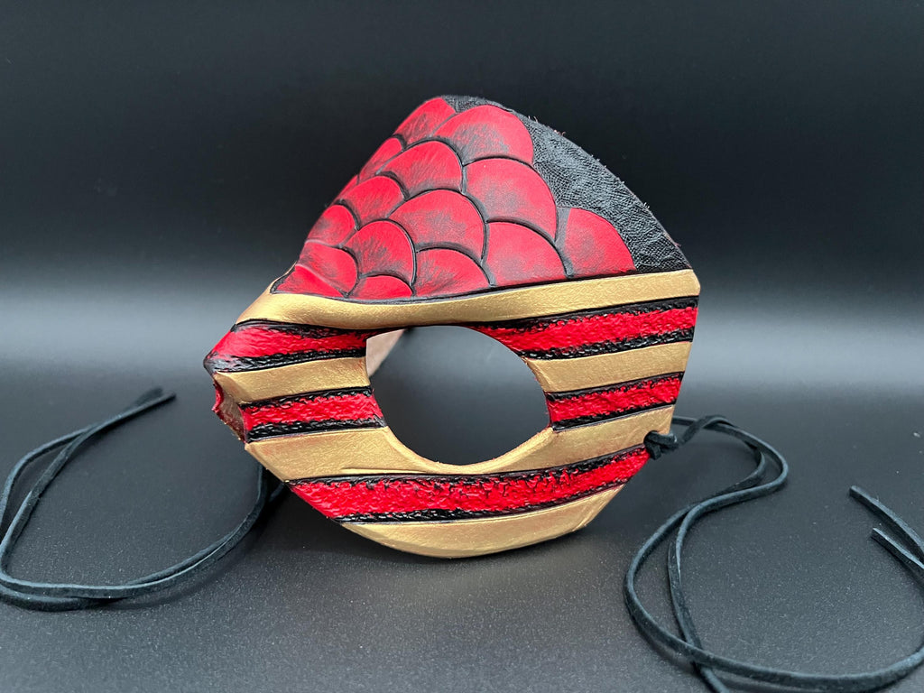 A leather mask featuring a red and black scales design with gold trim dragon motif shown on a dark gray background with black ties