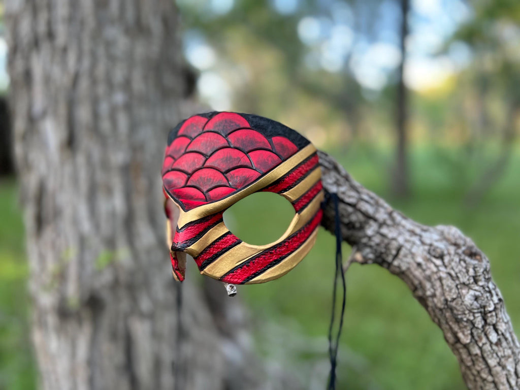 A handcrafted masquerade mask with a red and black scaled pattern and gold stripes, hanging from a tree branch in an outdoor park setting.
