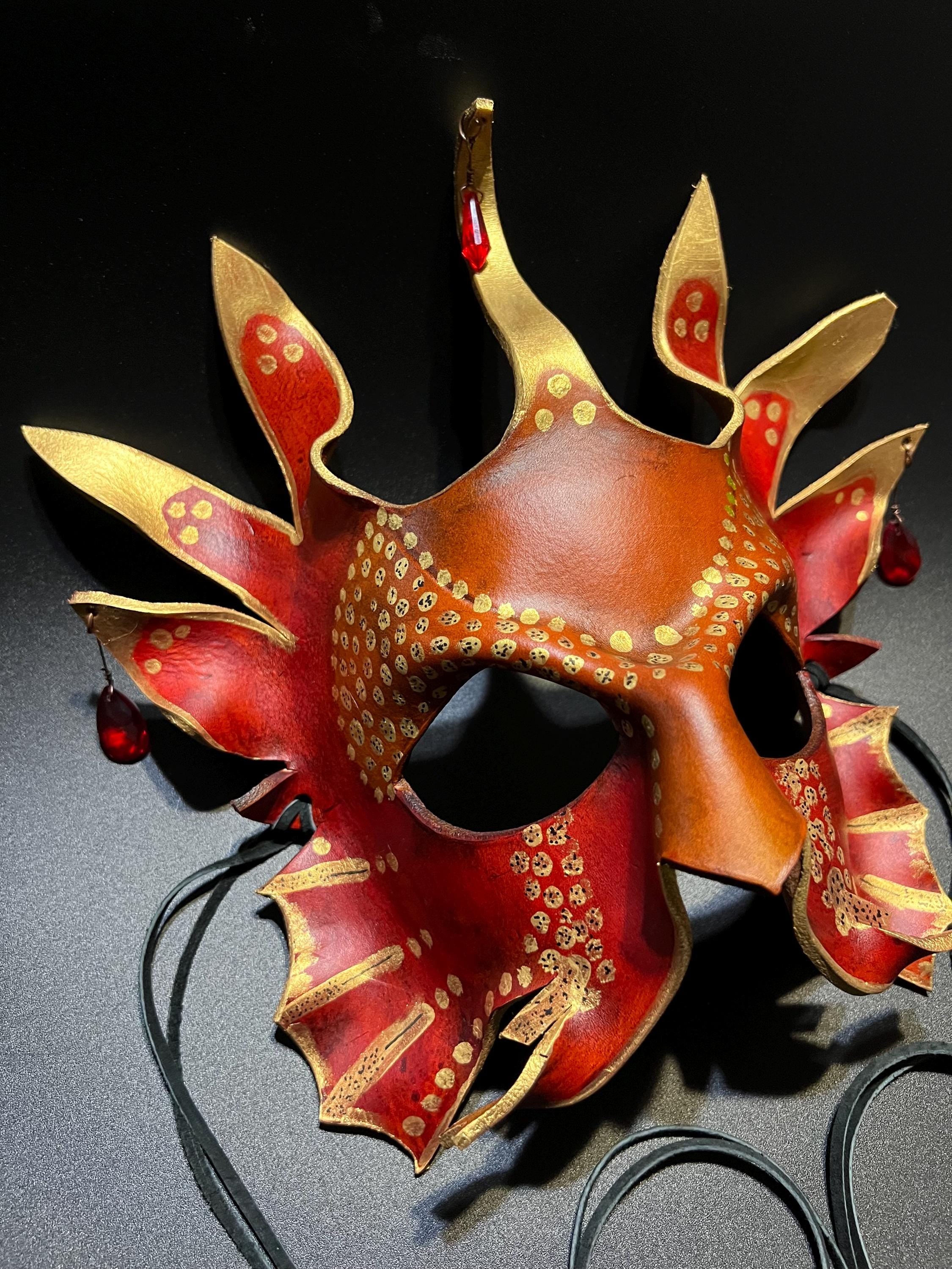 A decorative fantasy-style masquerade mask in reddish-brown leather with gold painted details, red accents, and black eye openings, photographed against a dark background. Black cords are visible behind it.