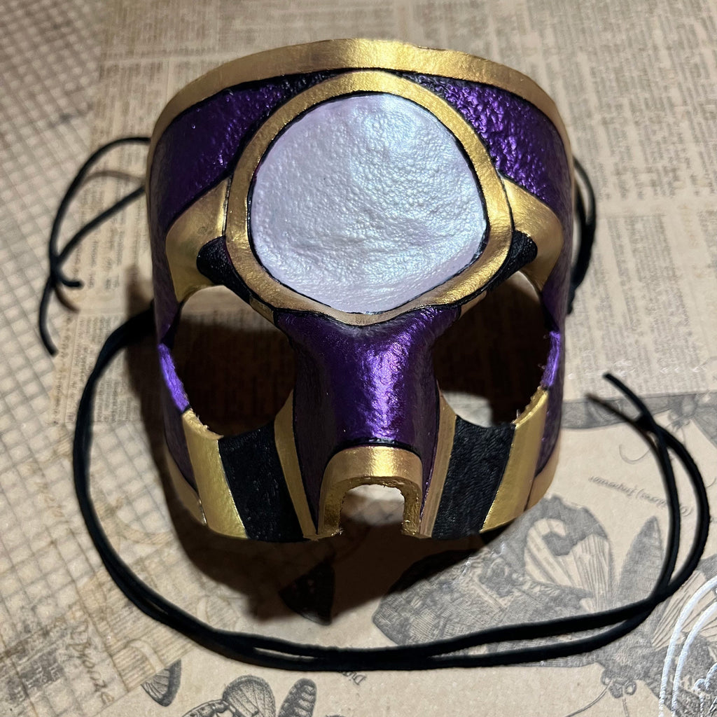 A handcrafted fantasy mask with a metallic purple and gold finish, featuring a large textured white circular eye detail and black ear loops, resting on a vintage book page background.
