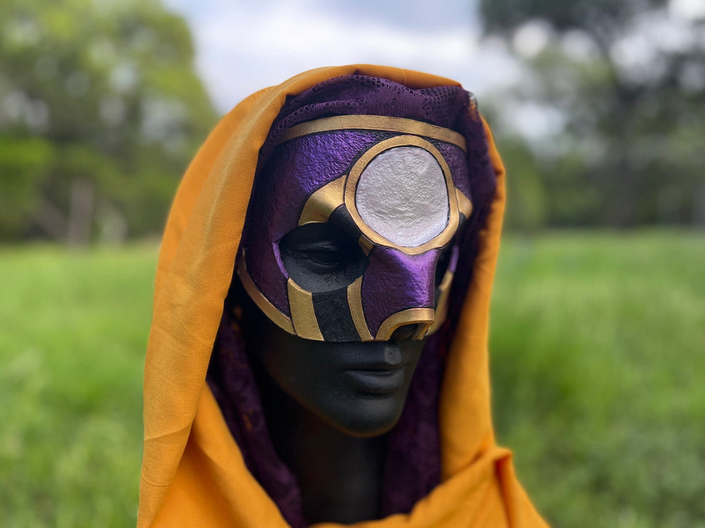 A person outdoors wearing a purple and gold mask with a matching purple hood and yellow-orange fabric draped over the shoulders, against a blurred green field background.