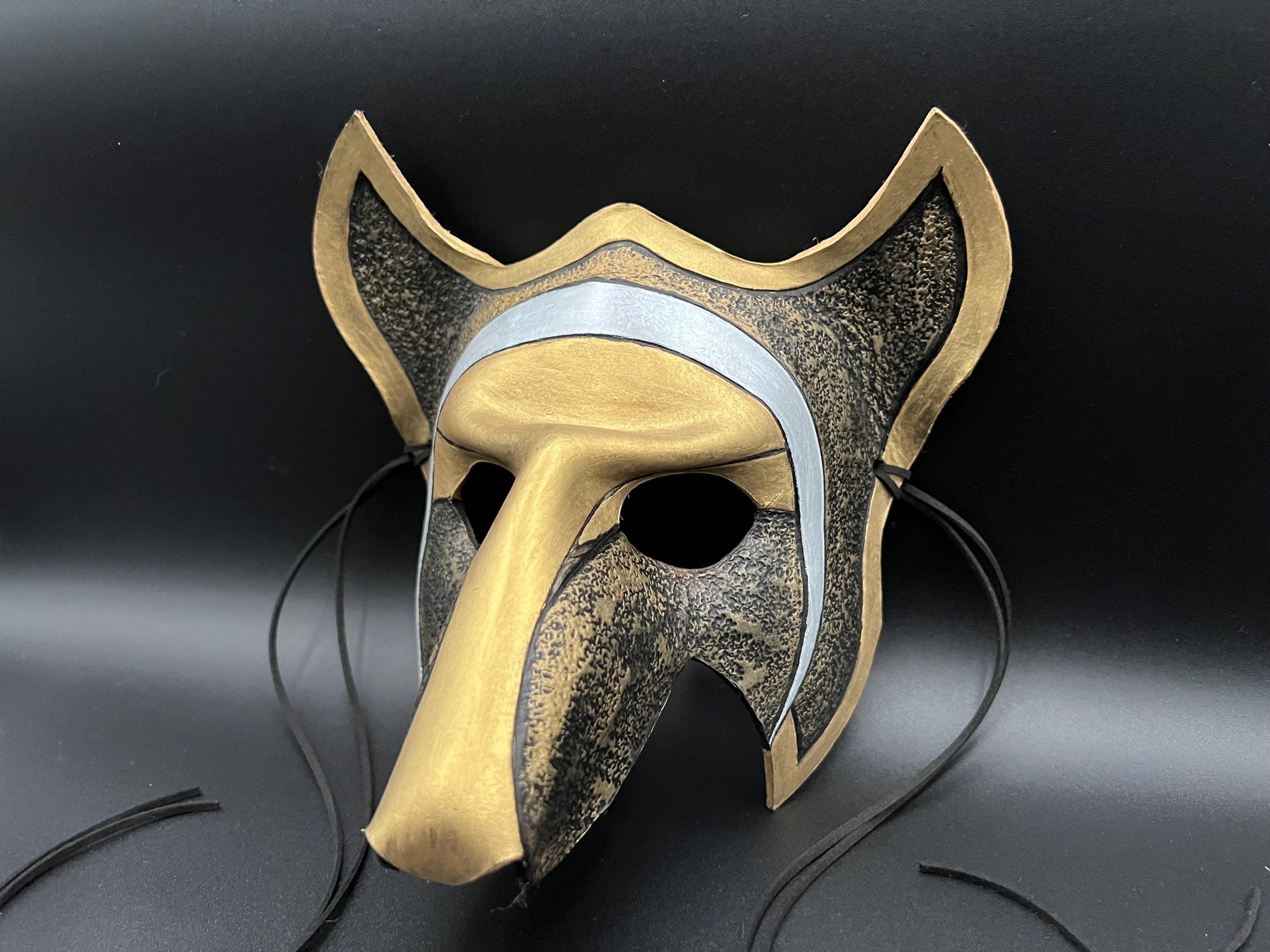 A stylized animal mask with a long snout, pointed ears, and black hanging straps, shown on a dark studio background. The mask has a metallic gold finish with a textured dark gray/black patterned section and a light silver band across the forehead.