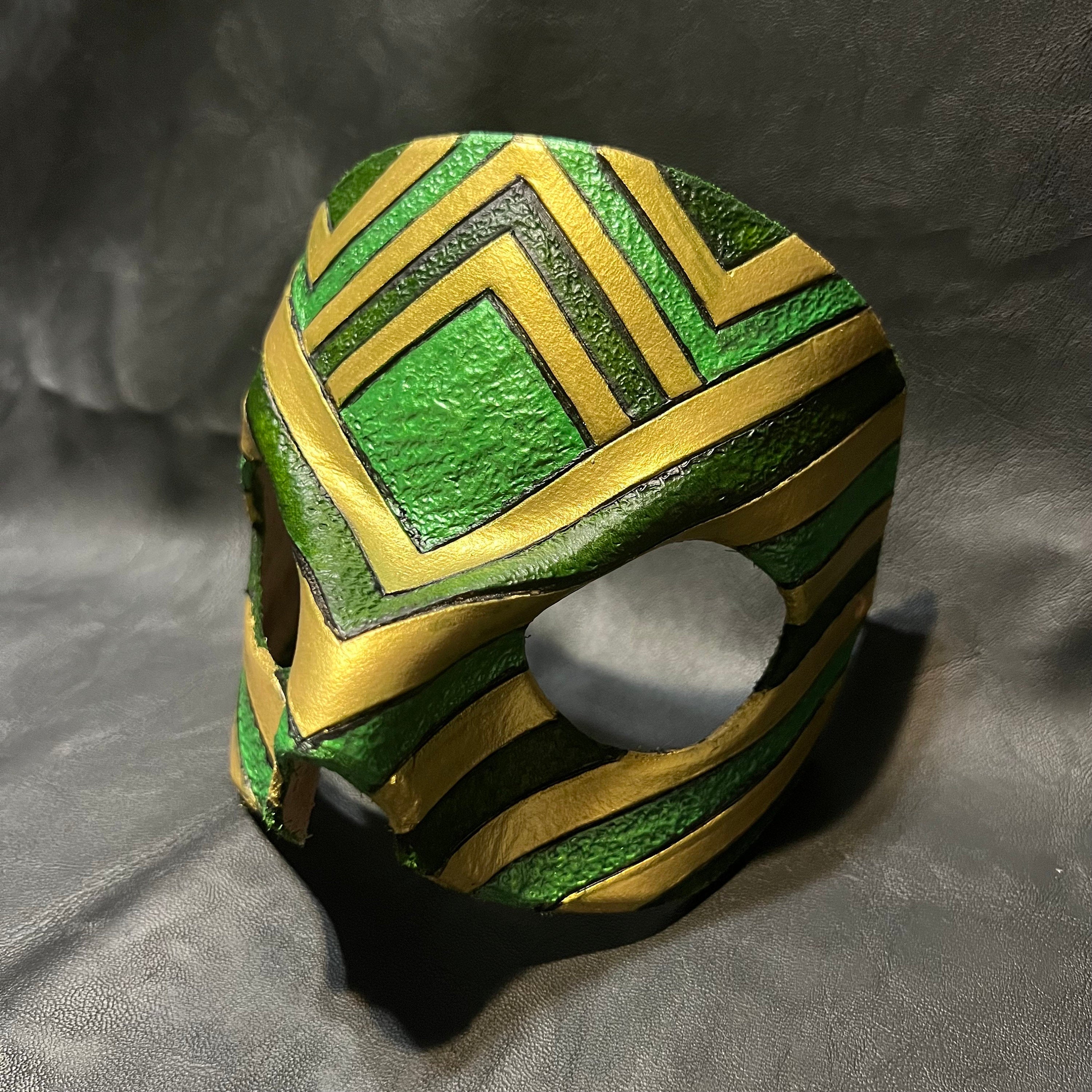 A green and gold lucha-style wrestling mask with geometric chevron patterns, photographed on a dark gray textured fabric background.