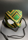 A green and gold patterned masquerade mask with black ear loops, displayed on a gray surface against a dark gray background.