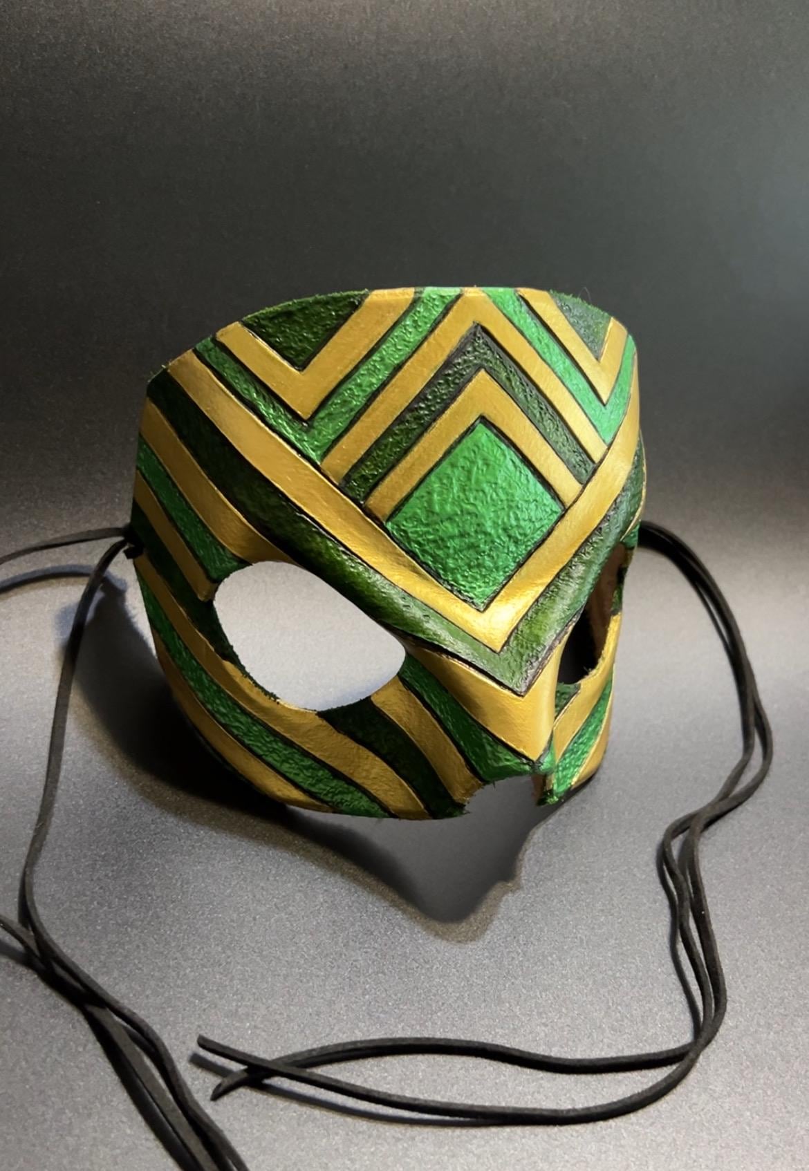 A green and gold patterned masquerade mask with black ear loops, displayed on a gray surface against a dark gray background.