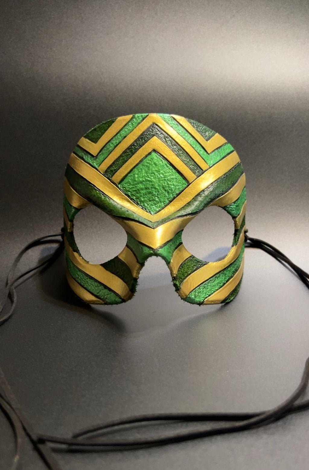 A green and gold patterned masquerade mask with eye cutouts and black ties, displayed on a dark gray background.