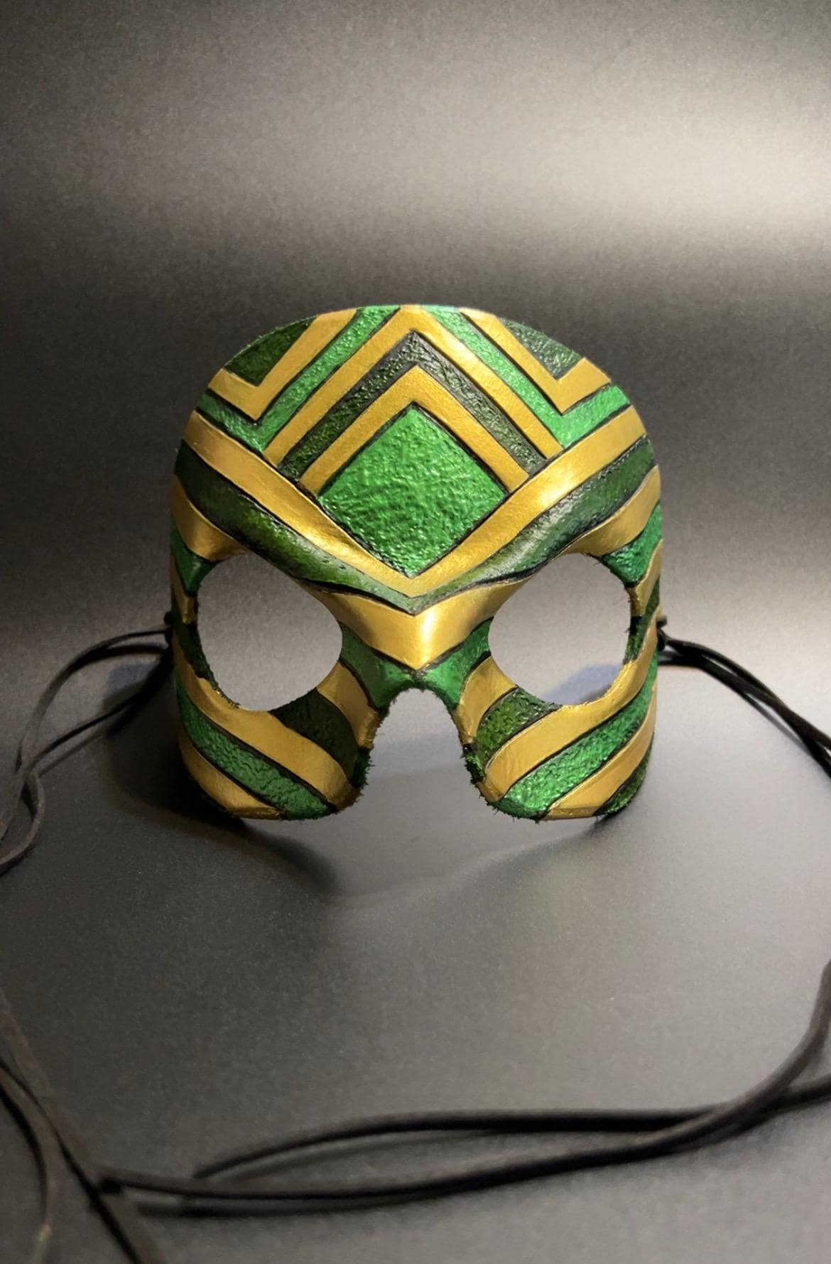 A green and gold patterned masquerade mask with eye cutouts and black ties, displayed on a dark gray background.
