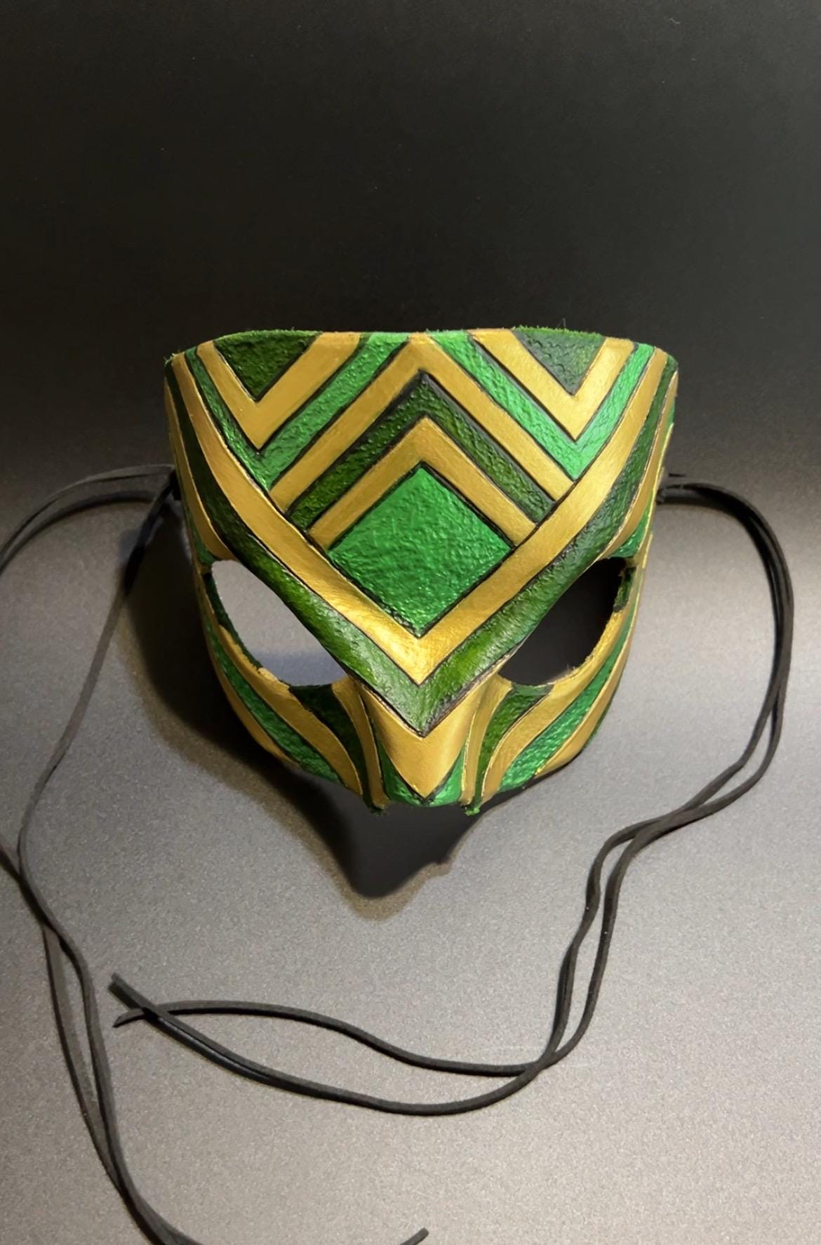 A green and gold patterned leather mask with black ties, displayed on a dark gray surface against a dark background.