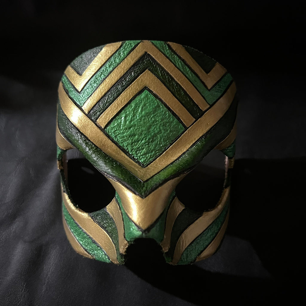 A green and gold patterned leather mask with eye cutouts, photographed against a dark black fabric background.