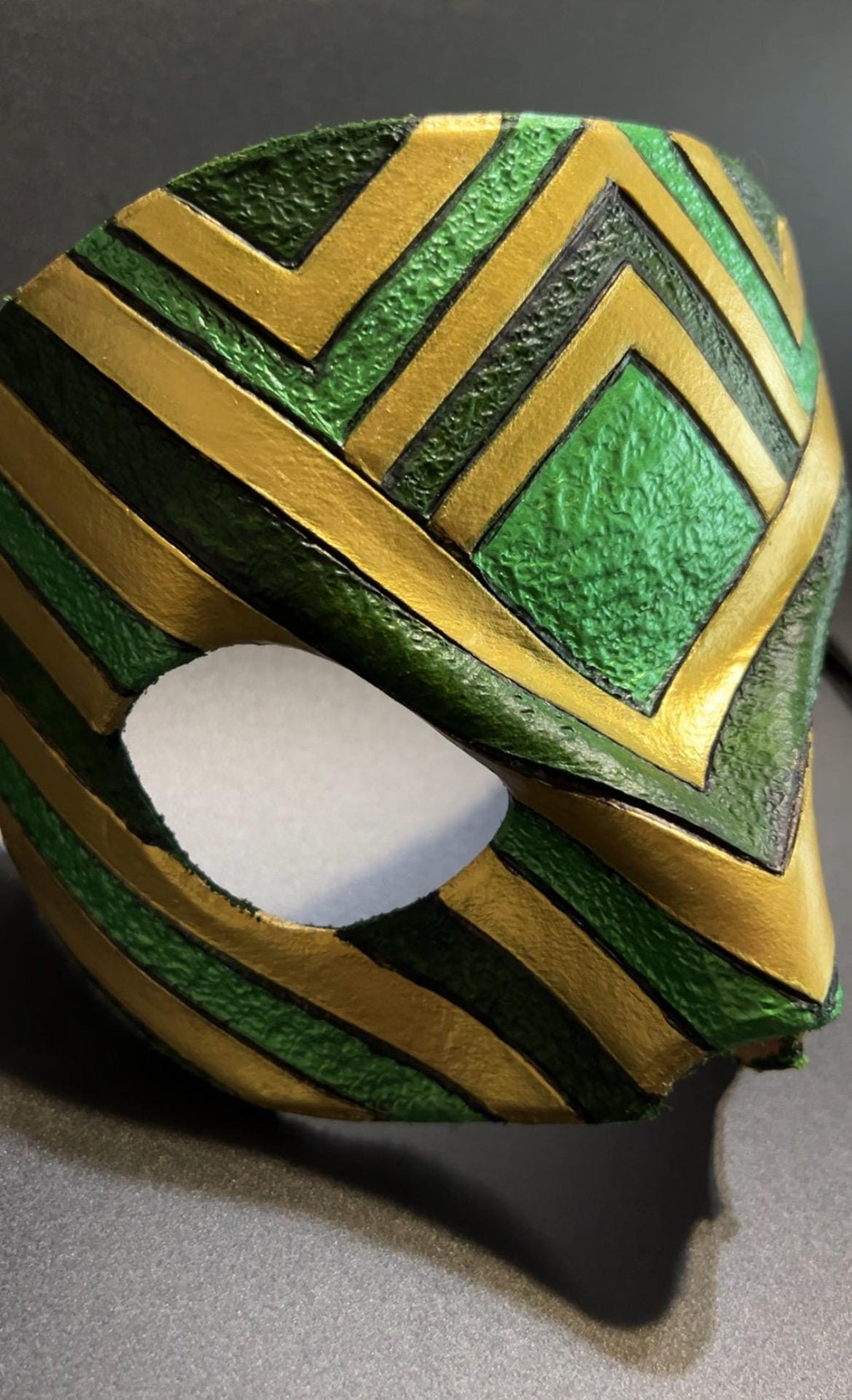 A close-up of a textured green and gold masquerade mask with an eye opening, resting on a dark gray surface.