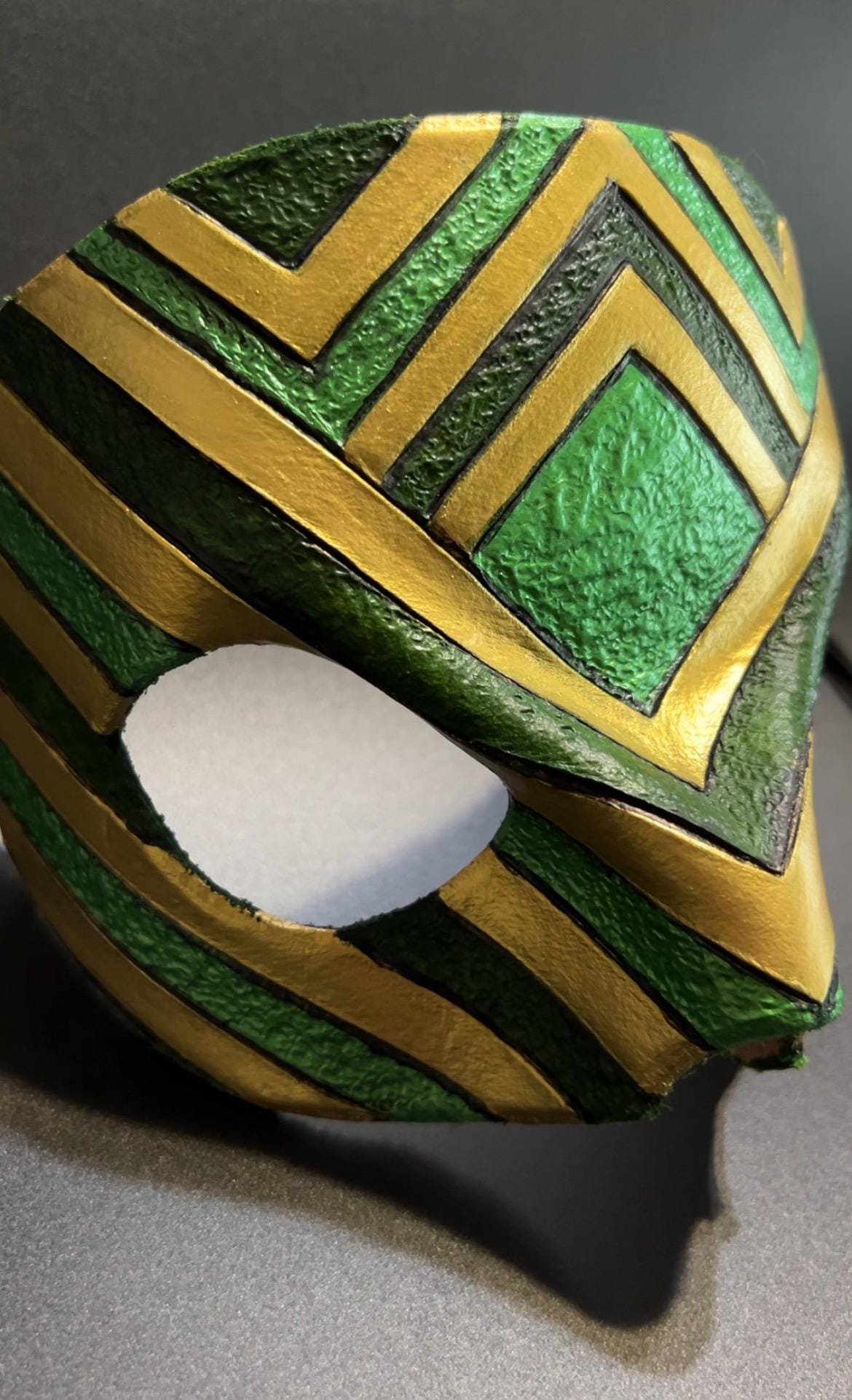 A close-up of a textured green and gold masquerade mask with an eye opening, resting on a dark gray surface.
