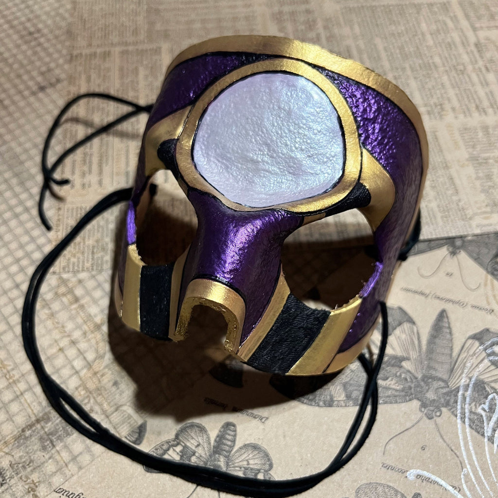 A purple and gold mask with a white circular center, resting on a vintage-style illustrated paper background with black suede cords.