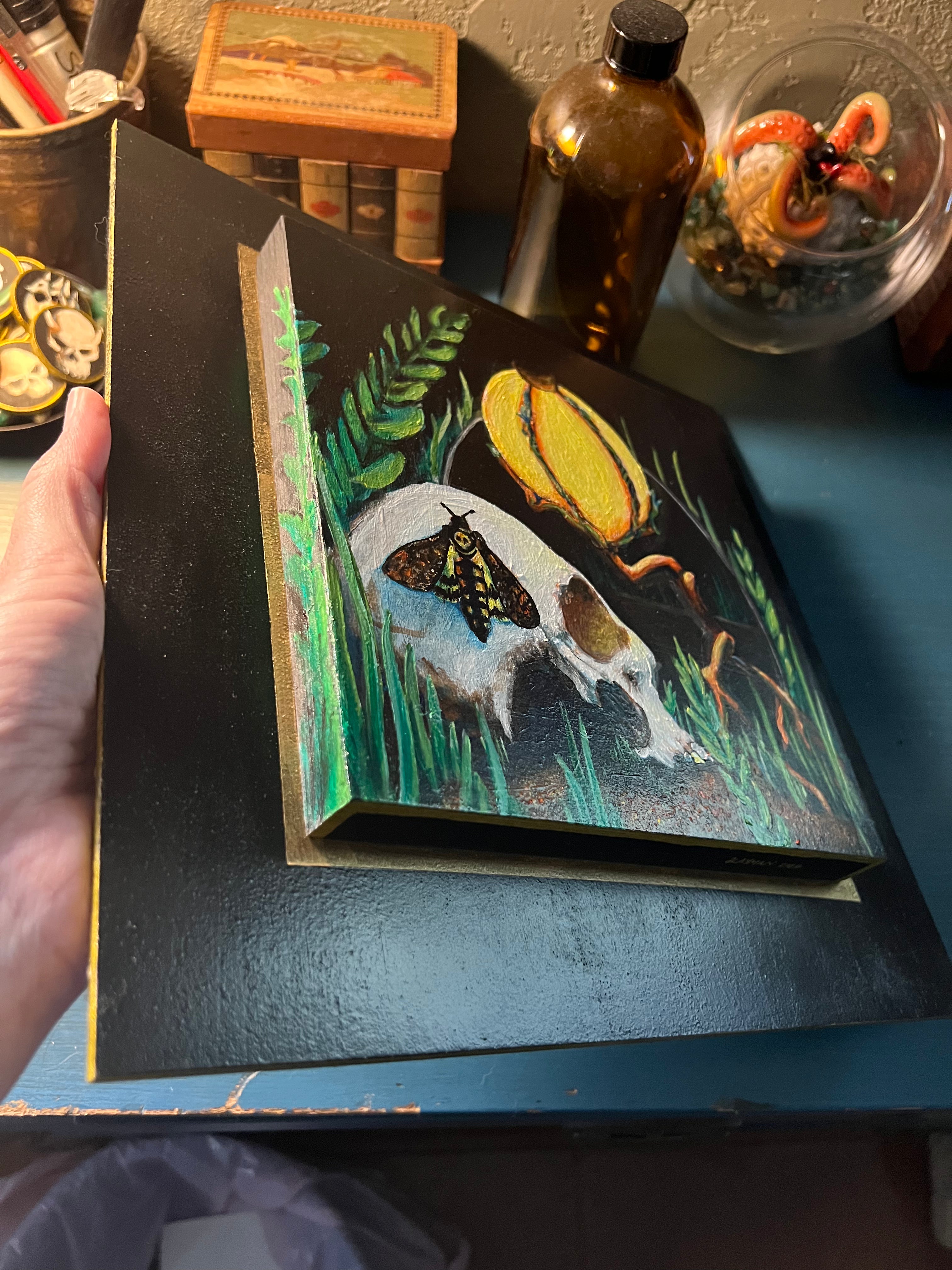 A hand holds a small black canvas painting featuring a white skull with a butterfly, green grass, and a yellow moon, displayed on a blue table with bottles and other objects in the background.