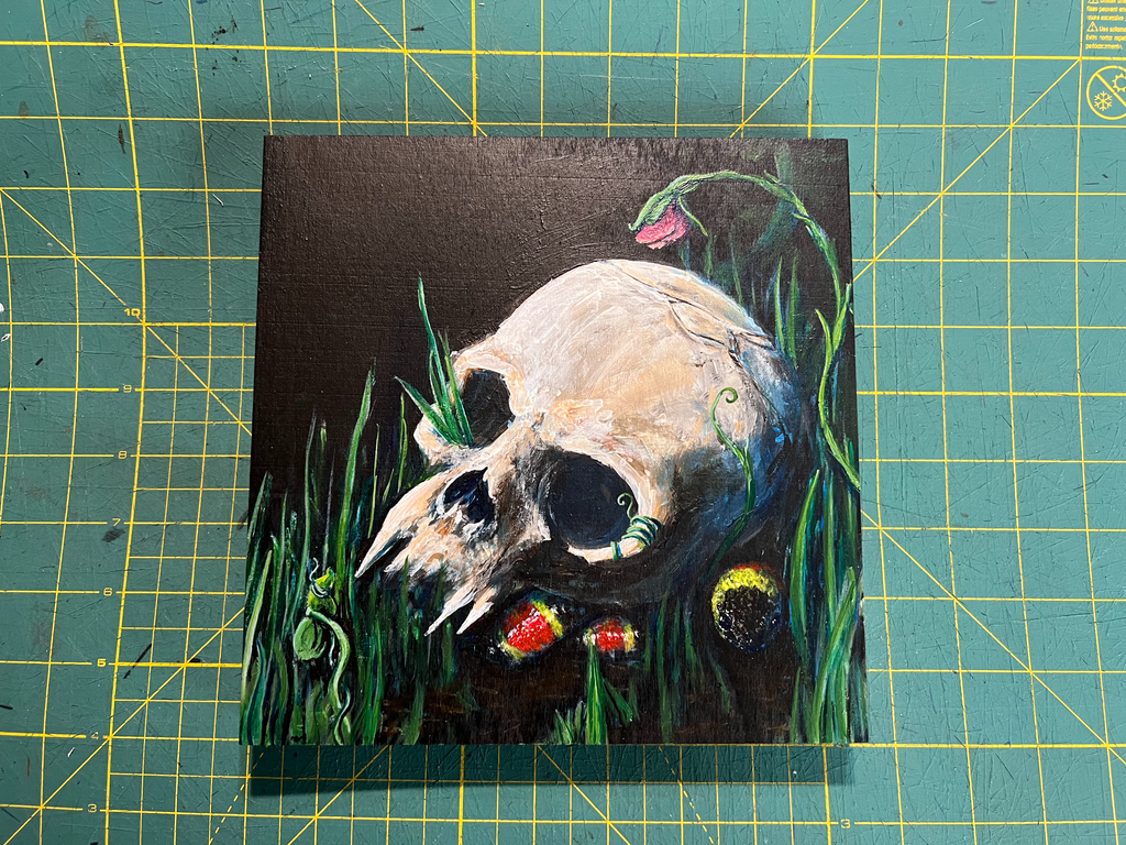 A square canvas painting of a human skull resting in green grass with small red and yellow flowers, set against a dark background, displayed on a green cutting mat with yellow grid lines.