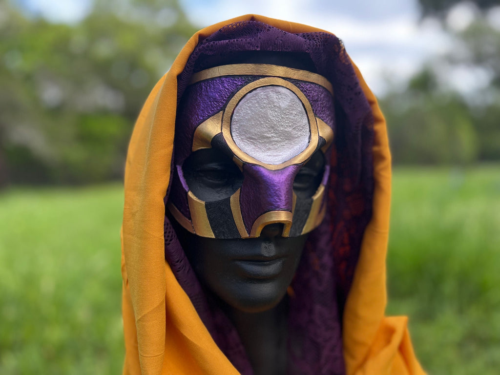 A hooded mask with a purple and gold design and a circular silver eye detail, displayed on a mannequin head outdoors with a blurred green field background.