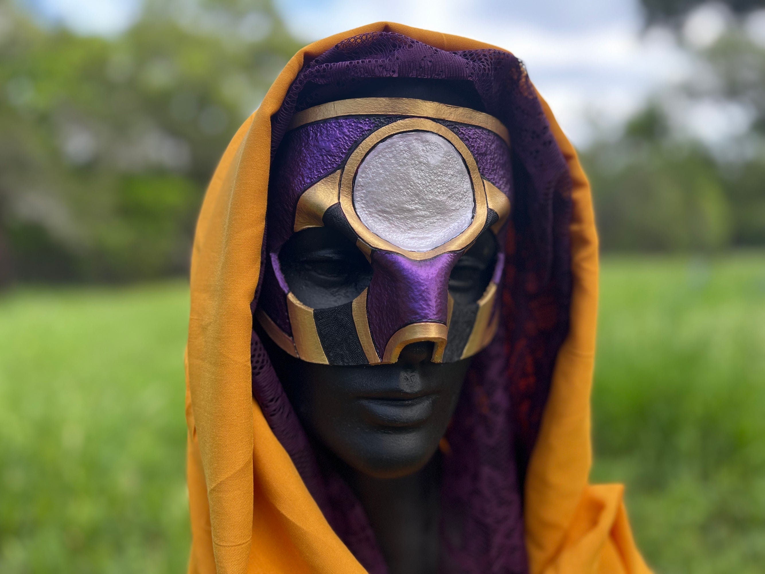 A hooded mask with a purple and gold design and a circular silver eye detail, displayed on a mannequin head outdoors with a blurred green field background.