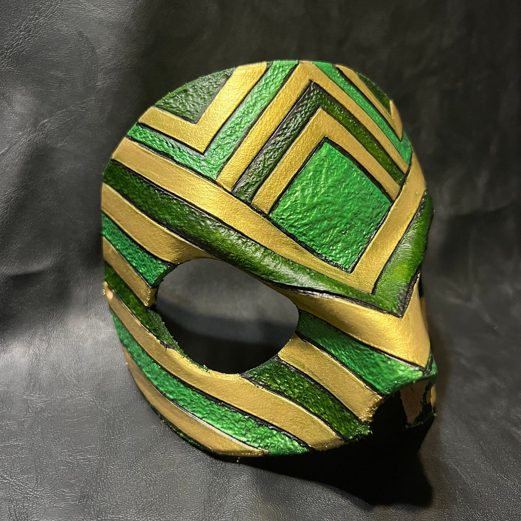 A green and gold patterned leather mask with a textured finish, photographed on a dark gray textured background.
