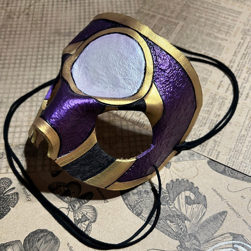 A purple and gold masquerade mask with a black strap, resting on a vintage-style illustrated paper background.
